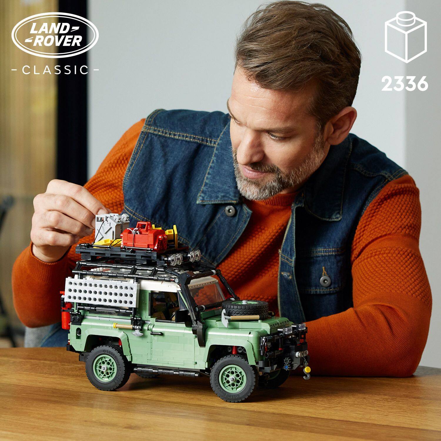The man is sitting at a table, working on a toy Land Rover Classic, which is a model of a classic Land Rover. He is carefully assembling the toy, which is made up of blocks and building sets. The toy is placed on the table, and the man is focused on putting it together.