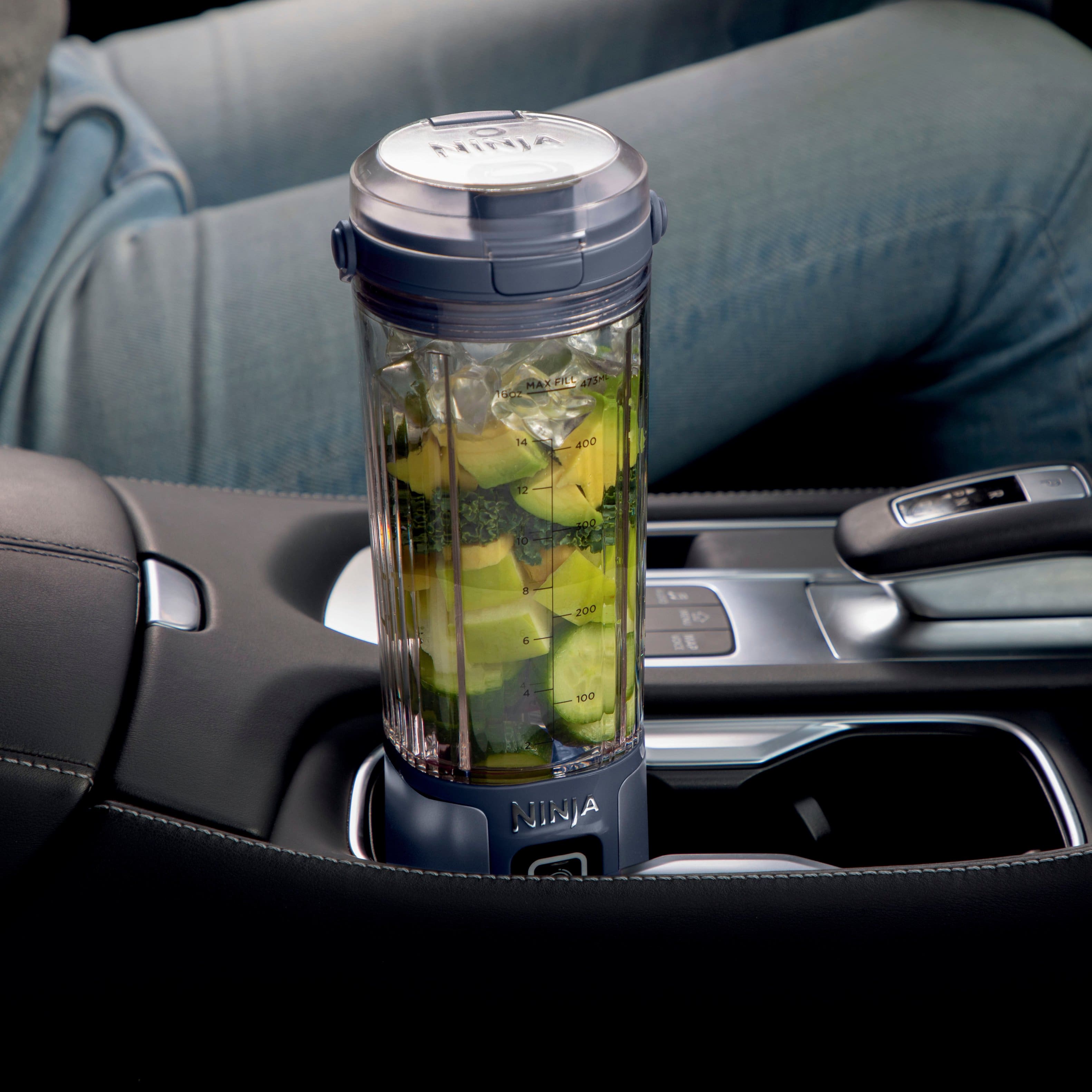 The image features a Ninja MaxFRL blender sitting in a car cup holder. The blender is filled with green vegetables, and it is placed in the cup holder, making it convenient for travel and portability. The cup holder is located in the car, and the blender is ready to be used for making smoothies or other blended beverages.