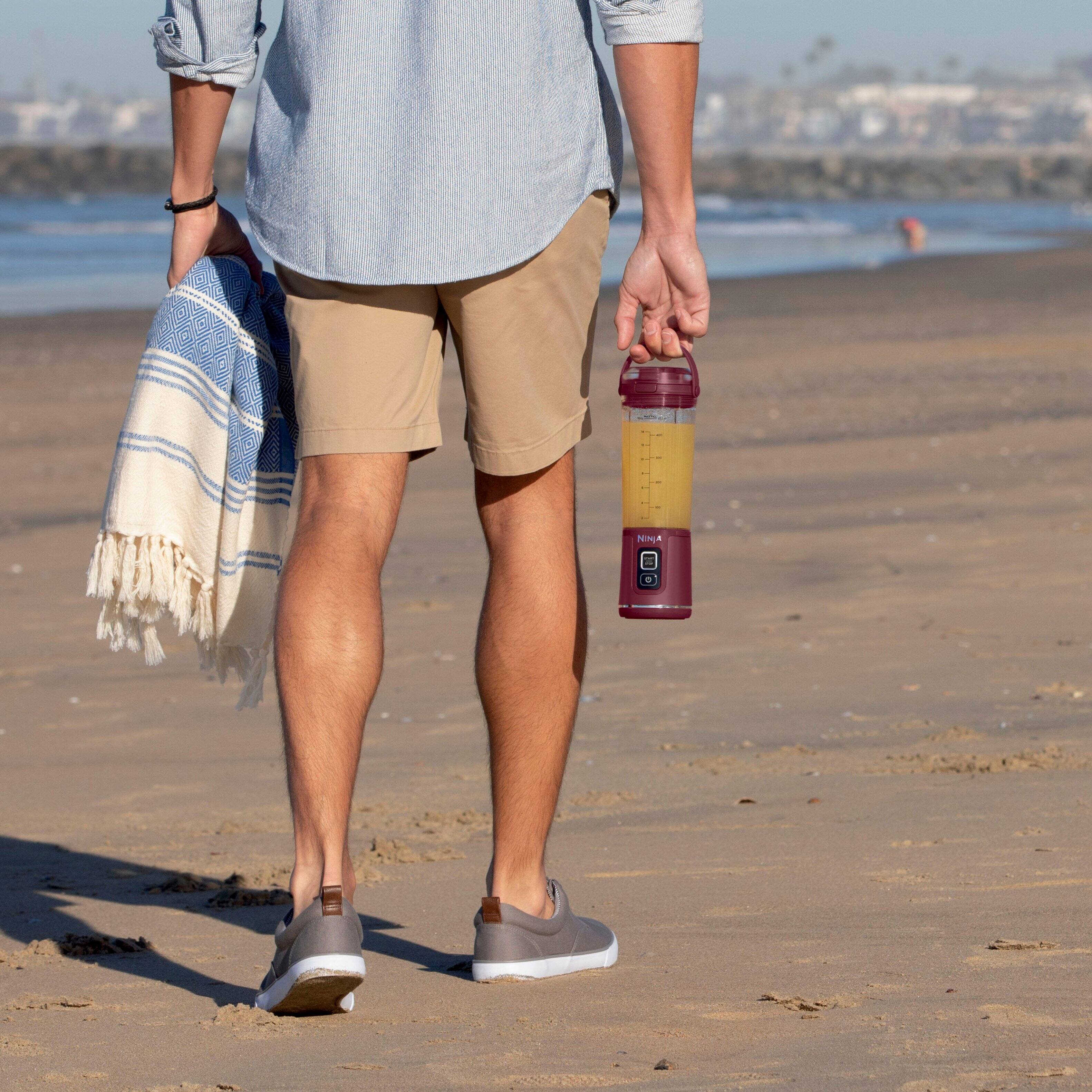The image features a man walking on a beach with a towel and a water bottle. The water bottle is described as "hyper portable" and has a hinged carry handle for easy transportation. The towel is also being carried by the man, making it a convenient and practical accessory for a day at the beach.