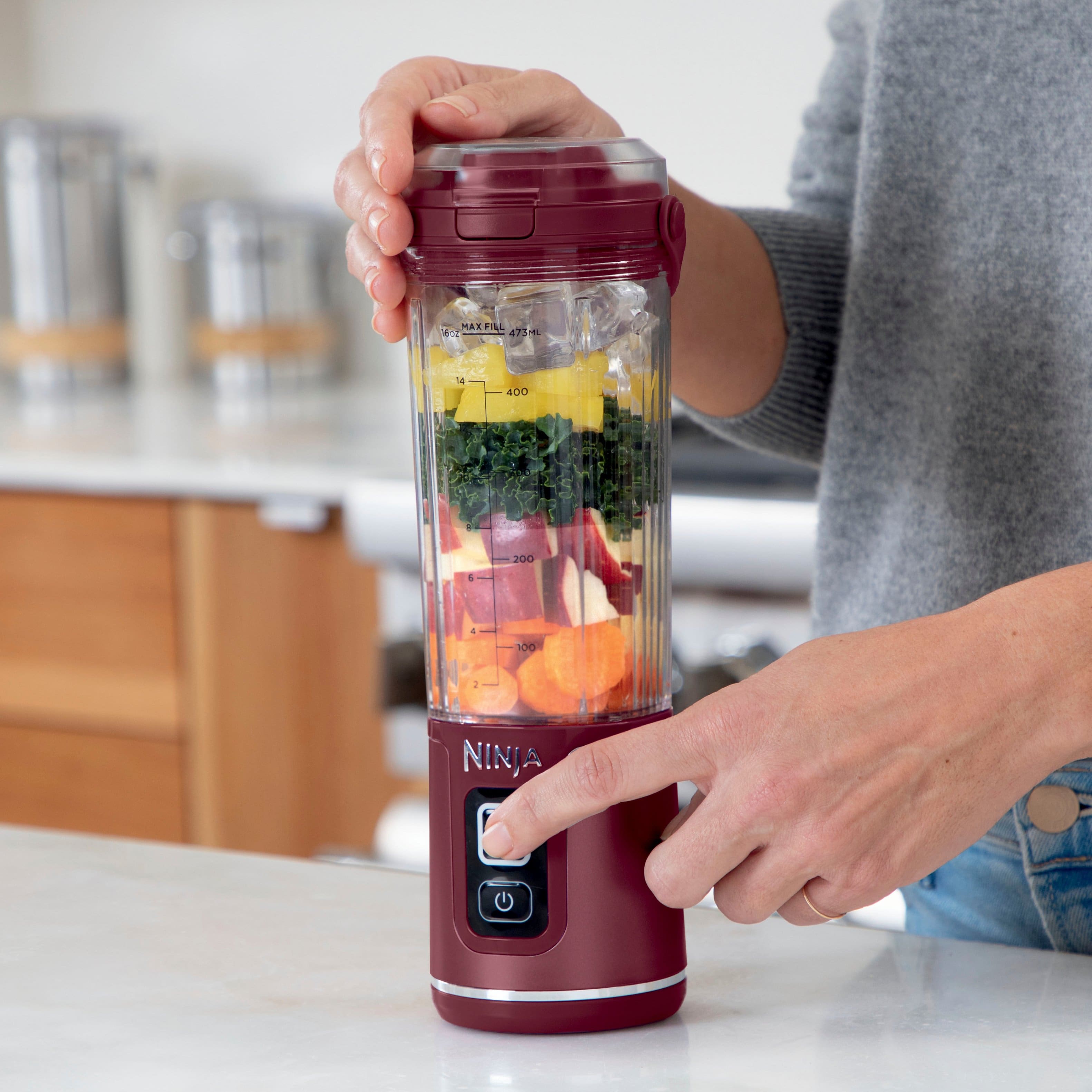 The image shows a person holding a red Ninja blender filled with fruits and vegetables. The blender is easy to use with separate power and blend buttons, ensuring no unintended blending. The person is standing in a kitchen, surrounded by various items such as a bowl, a sink, and a refrigerator. The scene suggests that the person is preparing a healthy and nutritious meal using the Ninja blender.