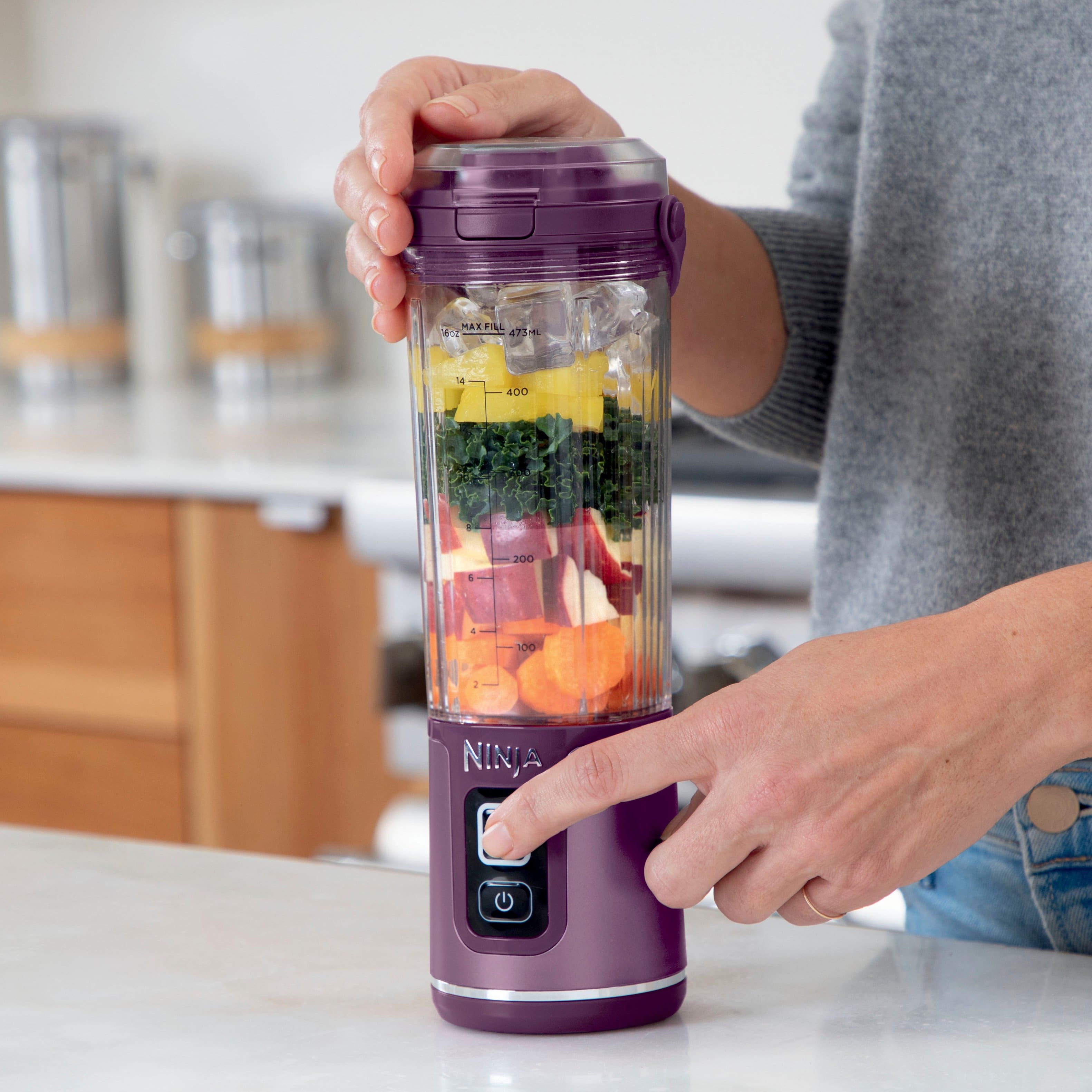 The image features a person holding a Ninja blender with a variety of fruits and vegetables inside. The blender is easy to use with separate power and blend buttons, ensuring no unintended blending. The person is standing in a kitchen, and the blender is placed on a countertop.