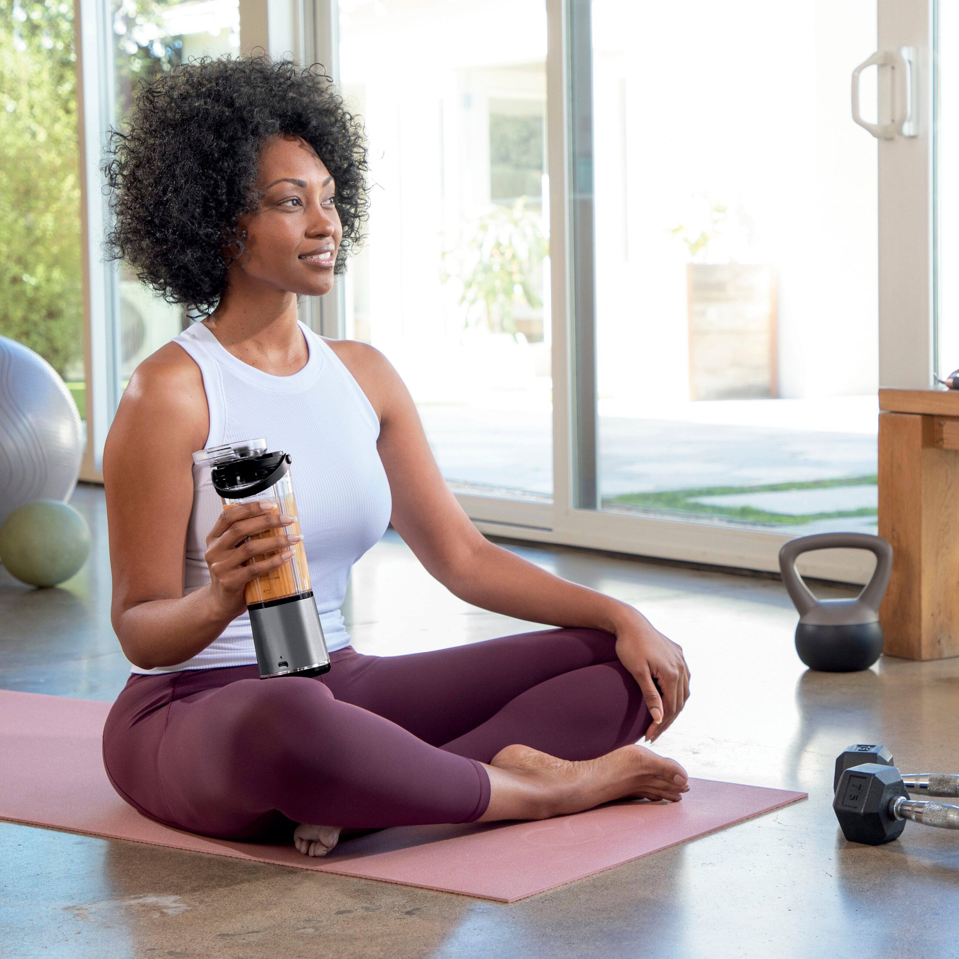 The image shows a woman sitting on the floor, holding a blender. The blender is placed on the floor next to her. The woman is smiling, indicating that she is enjoying the moment. The scene suggests that she might be preparing a smoothie or a blended drink using the blender.