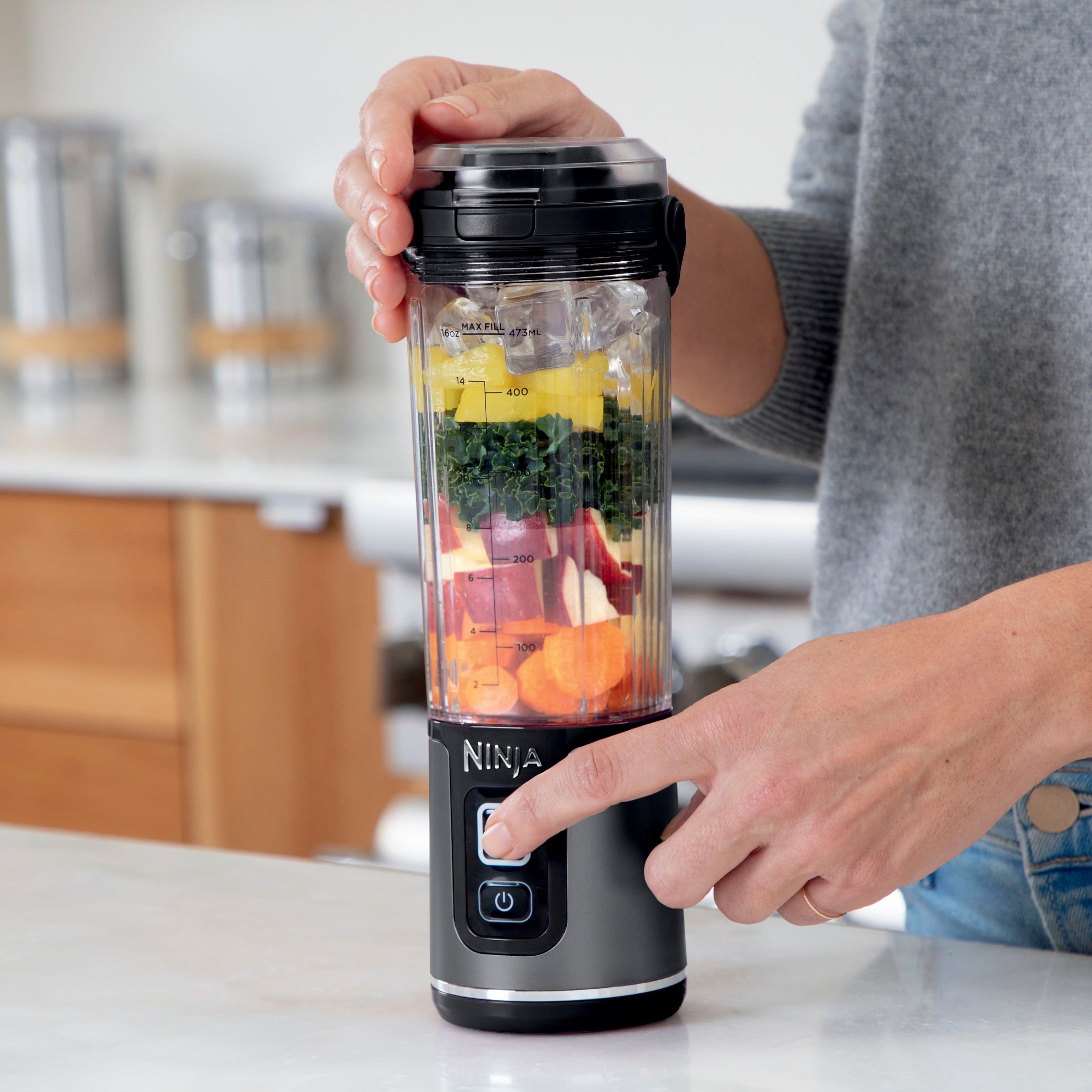 The image features a person holding a Ninja blender filled with fruits and vegetables. The blender is easy to use with separate power and blend buttons, ensuring that there is no unintended blending. The person is likely preparing a nutritious smoothie or juice using the blender's contents.