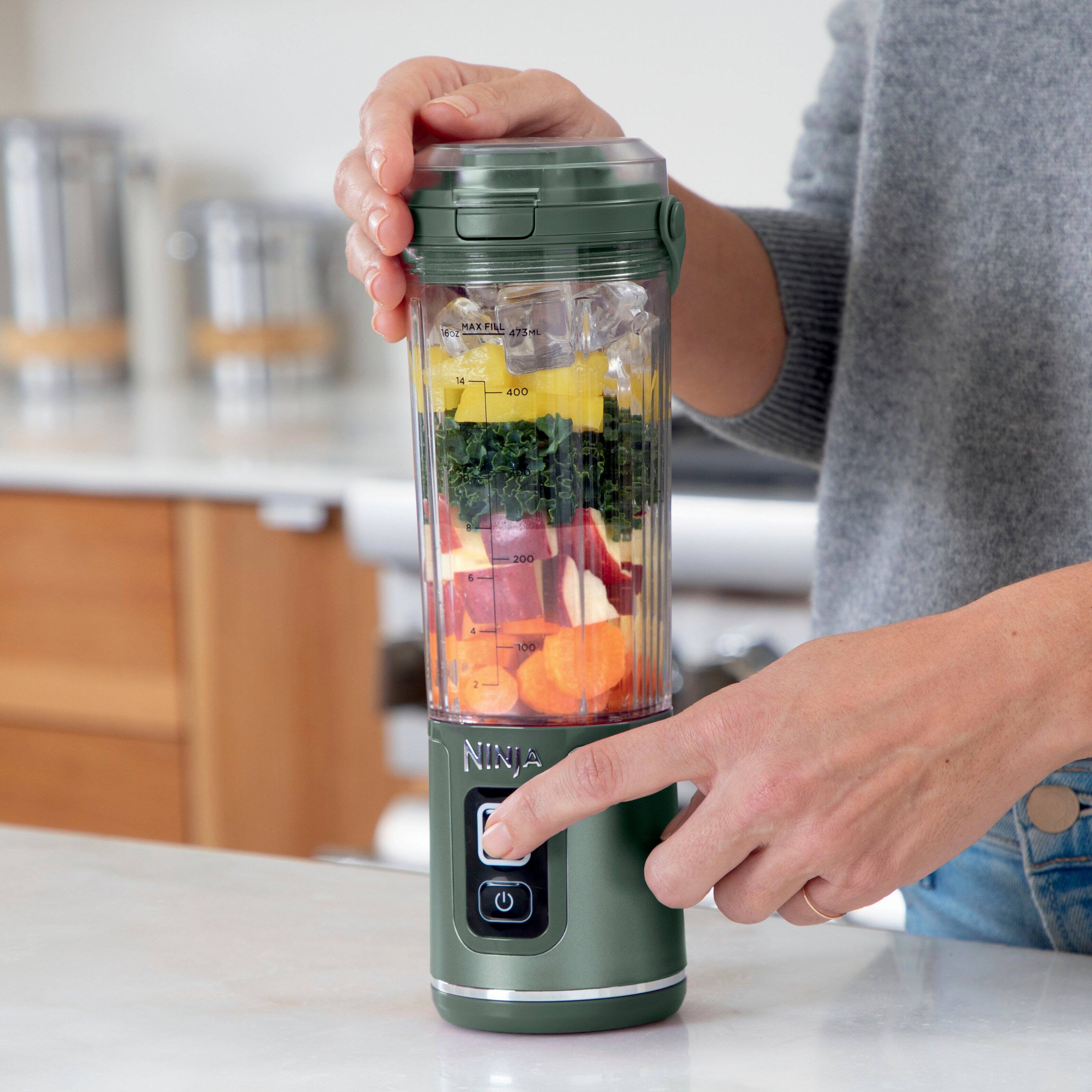 The image features a person holding a Ninja blender, which is easy to use with separate power and blend buttons. This ensures that there is no unintended blending. The blender is filled with various fruits and vegetables, indicating that it is being used for blending or preparing a smoothie or a similar beverage.