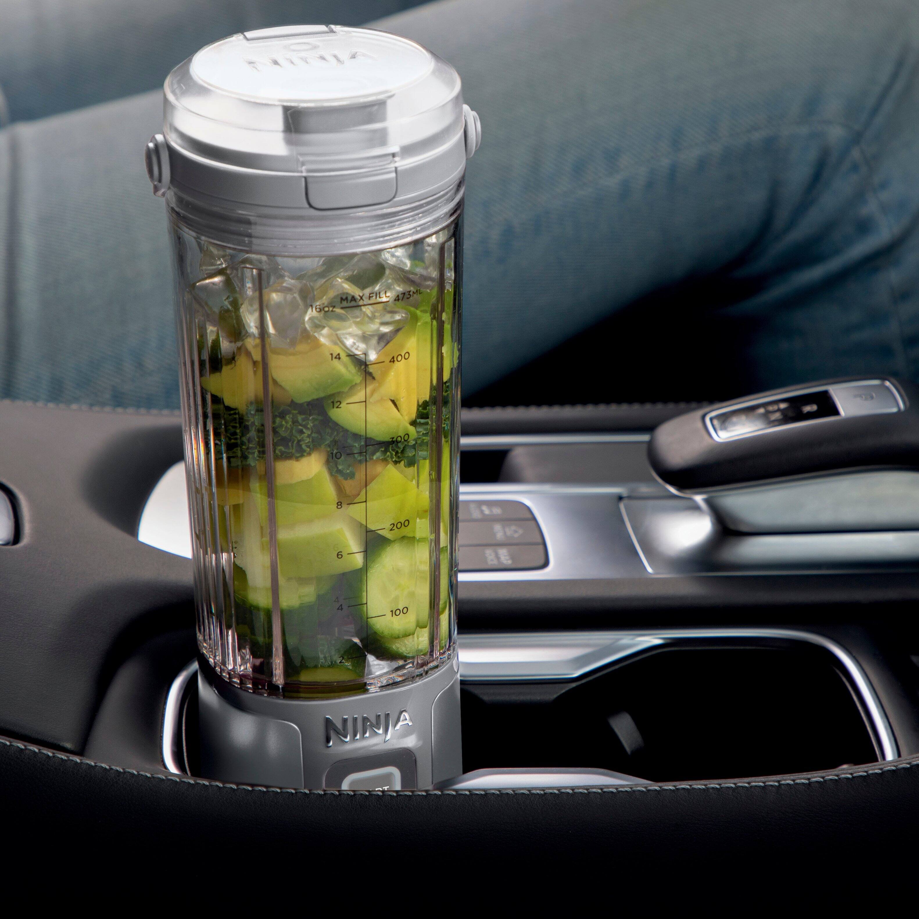The image features a car cup holder with a Ninja blender filled with green vegetables, such as broccoli and cucumbers. The cup holder is placed on a dashboard, and the blender is sitting on top of it. The scene suggests that the person who owns the car is using the cup holder as a convenient and portable way to store and blend their vegetables while on the go. The Ninja blender is known for its compact size and ease of use, making it a popular choice for those who want to have a healthy and nutritious snack or meal while traveling.