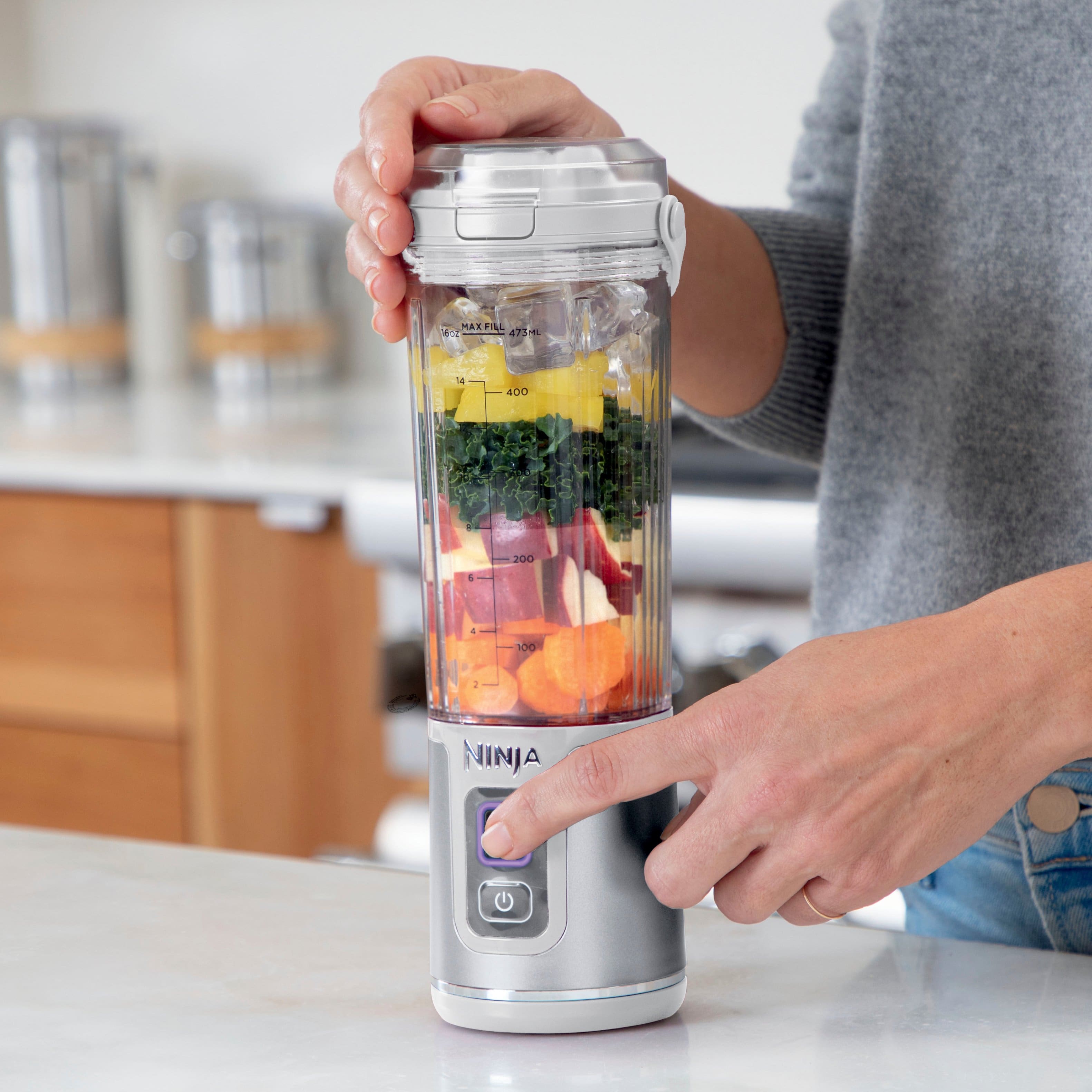 The image shows a person holding a Ninja blender with a variety of ingredients inside. The blender is filled with fruits and vegetables, making it easy to use with separate power and blend buttons. The person is demonstrating the blender's ease of use and the convenience of its design.