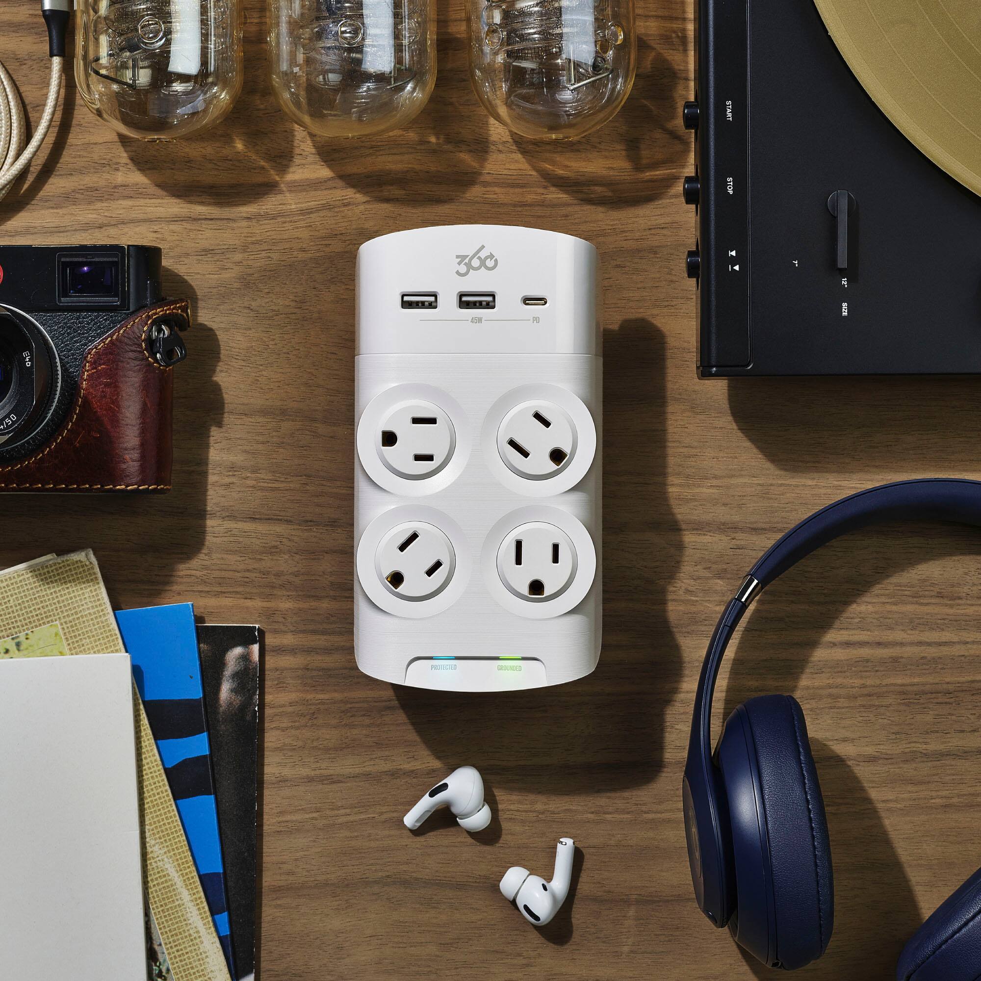 The image features a wooden table with various items on it, including a white 360 degree camera, a pair of earbuds, a camera, a record player, and a book. The table also has a white plug with four outlets on it, which is likely a surge protector. The arrangement of these items suggests that the table is a workspace or a place for leisure activities, such as photography, listening to music, and reading.