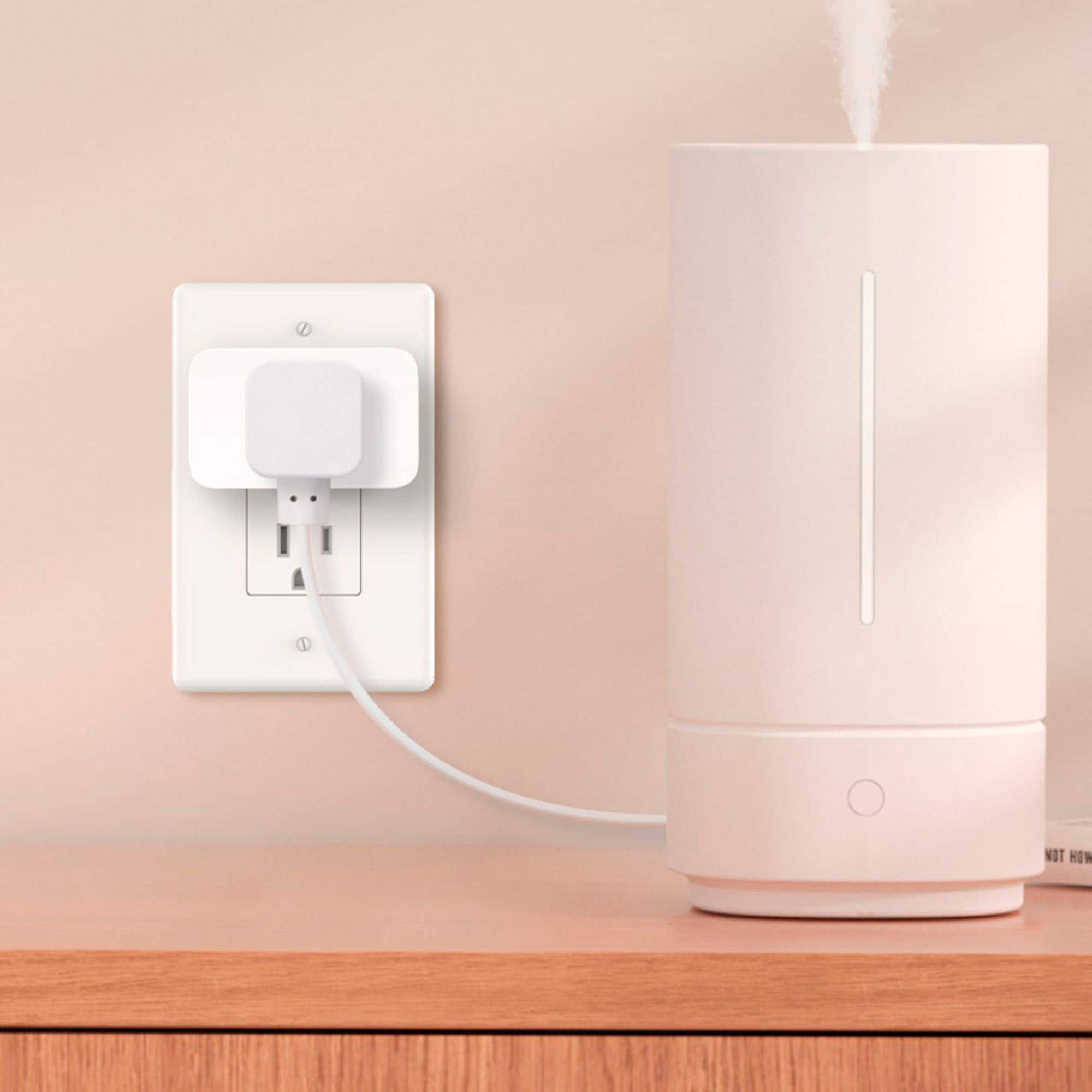 The image shows a white electrical appliance, likely a humidifier or air purifier, plugged into an outlet on a wooden desk. The appliance is connected to the outlet with a cord, and it is placed next to a wall. The appliance is designed to improve indoor air quality and maintain a comfortable humidity level in the room.
