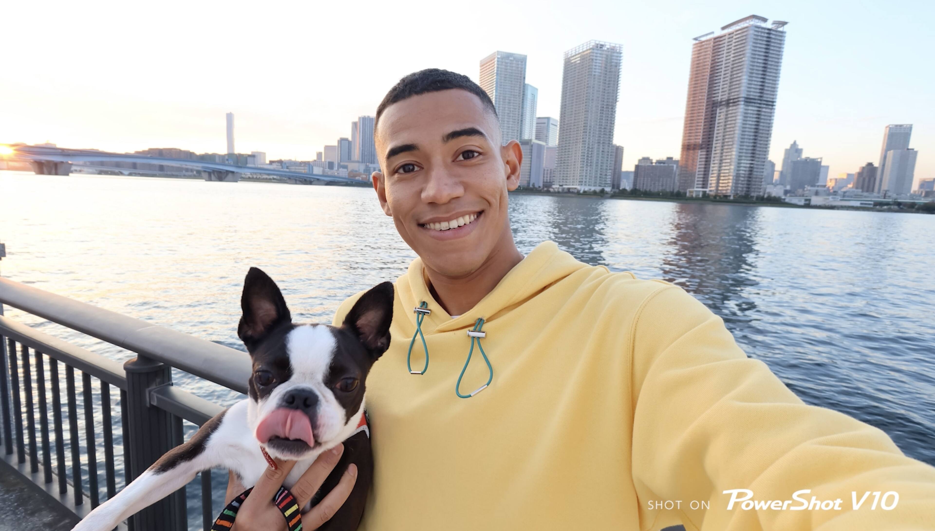 The image features a man wearing a yellow hoodie and holding a small dog. They are standing near a body of water, possibly a river or a lake. The man is smiling and appears to be enjoying the moment. The dog is also smiling, adding a playful and happy atmosphere to the scene. The image is captured using a PowerShot V10 camera, which is a digital camera known for its high-quality images and ease of use.