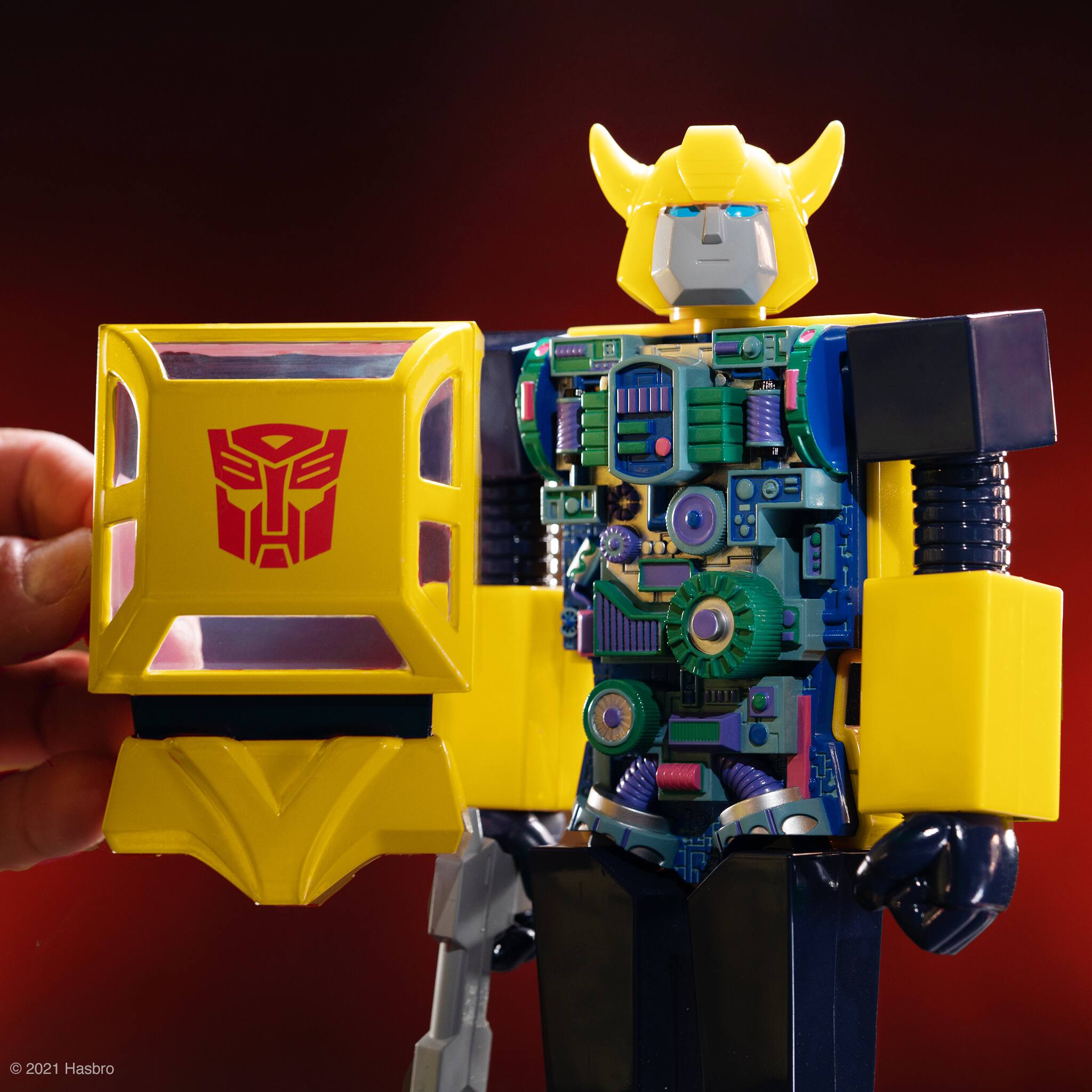 The image features a yellow and black action figure, possibly a Transformer or a robot, being held by a person. The action figure appears to be a toy, and it is being showcased in a red background. The figure is prominently displayed in the scene, capturing the viewer's attention.