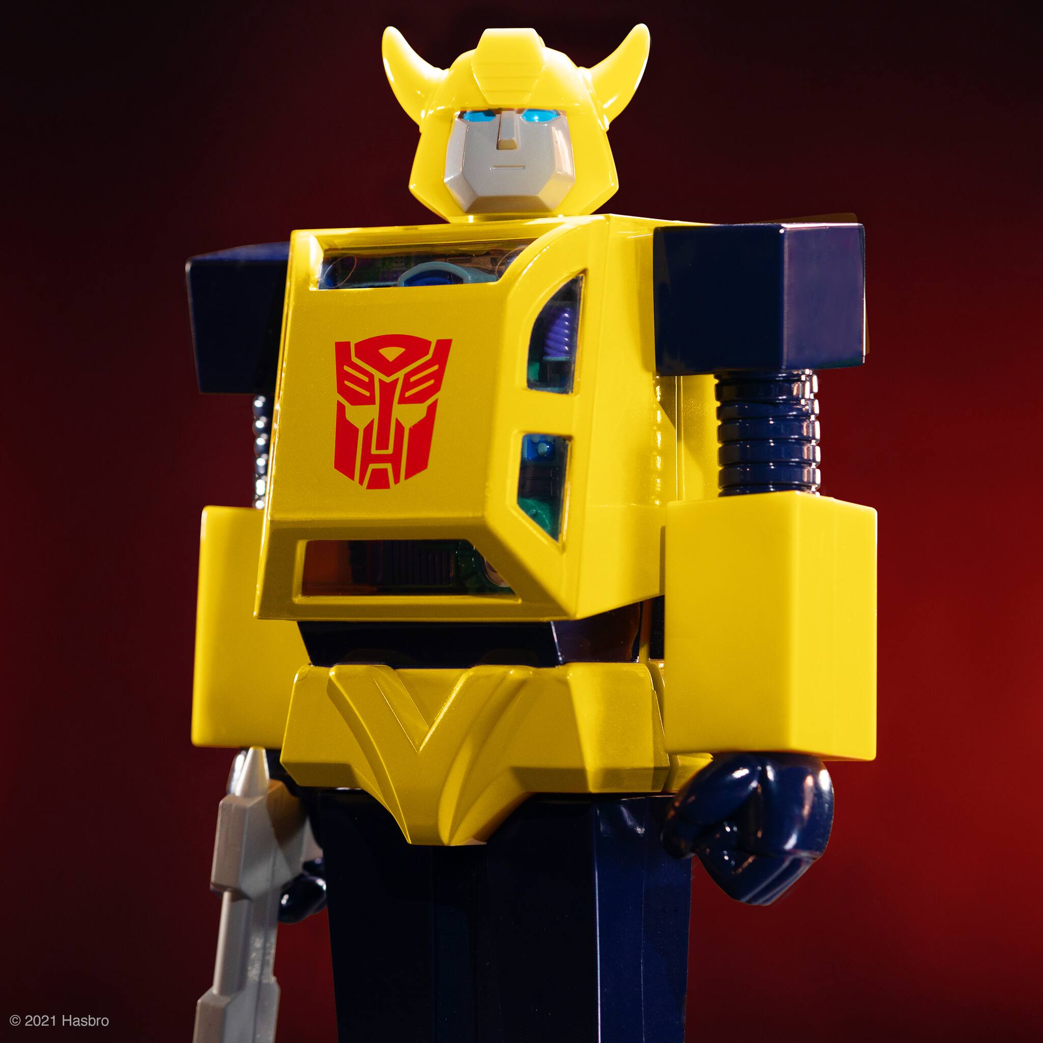 In the image, there is a yellow robot toy, possibly a Transformers figure, standing on a red background. The robot toy is wearing a yellow helmet and appears to be a part of a larger action figure collection. The toy is positioned in a way that it seems to be looking at the camera, giving it a more engaging and dynamic appearance.
