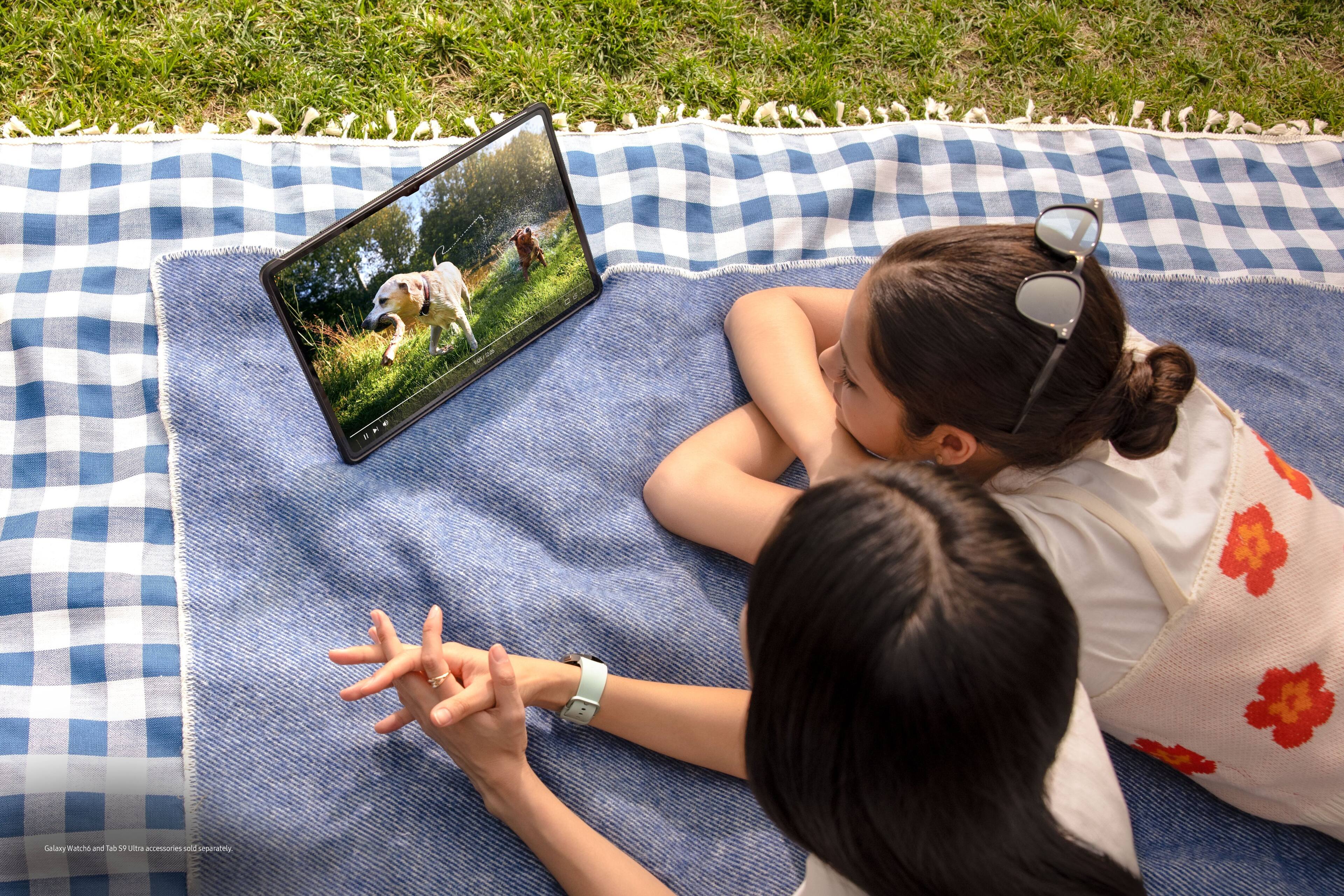 The image shows two girls sitting on a blanket outdoors, watching a video on a laptop. They are both focused on the screen, which is positioned in front of them. The laptop is placed on a checkered blanket, and the girls are sitting close to each other, enjoying their time together.