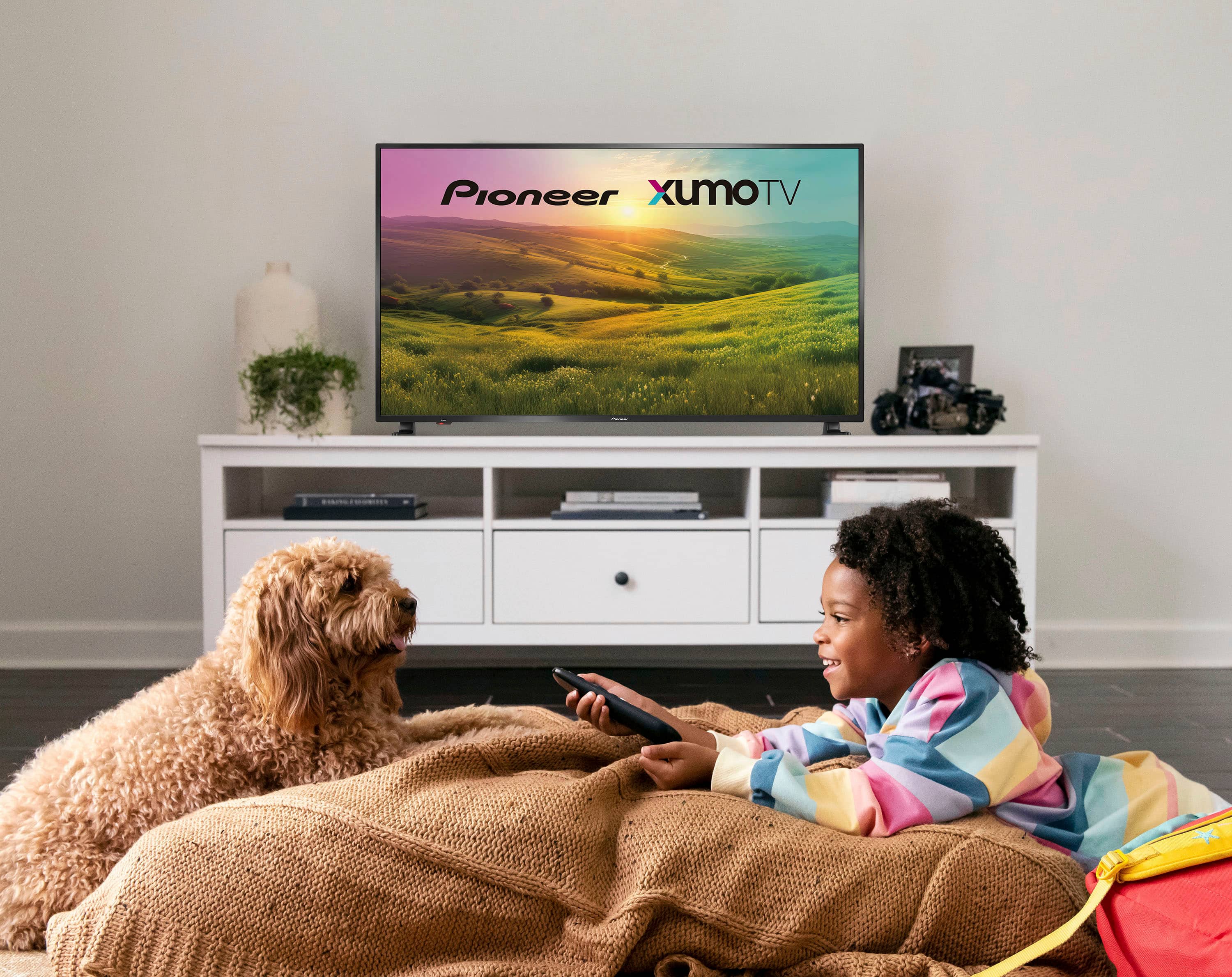 The image shows a young girl and a dog lying on a couch, watching television. The TV is a Pioneer XUMOTV, and the girl is holding a remote control in her hand. The scene appears to be a cozy and relaxing moment for both the girl and her dog.