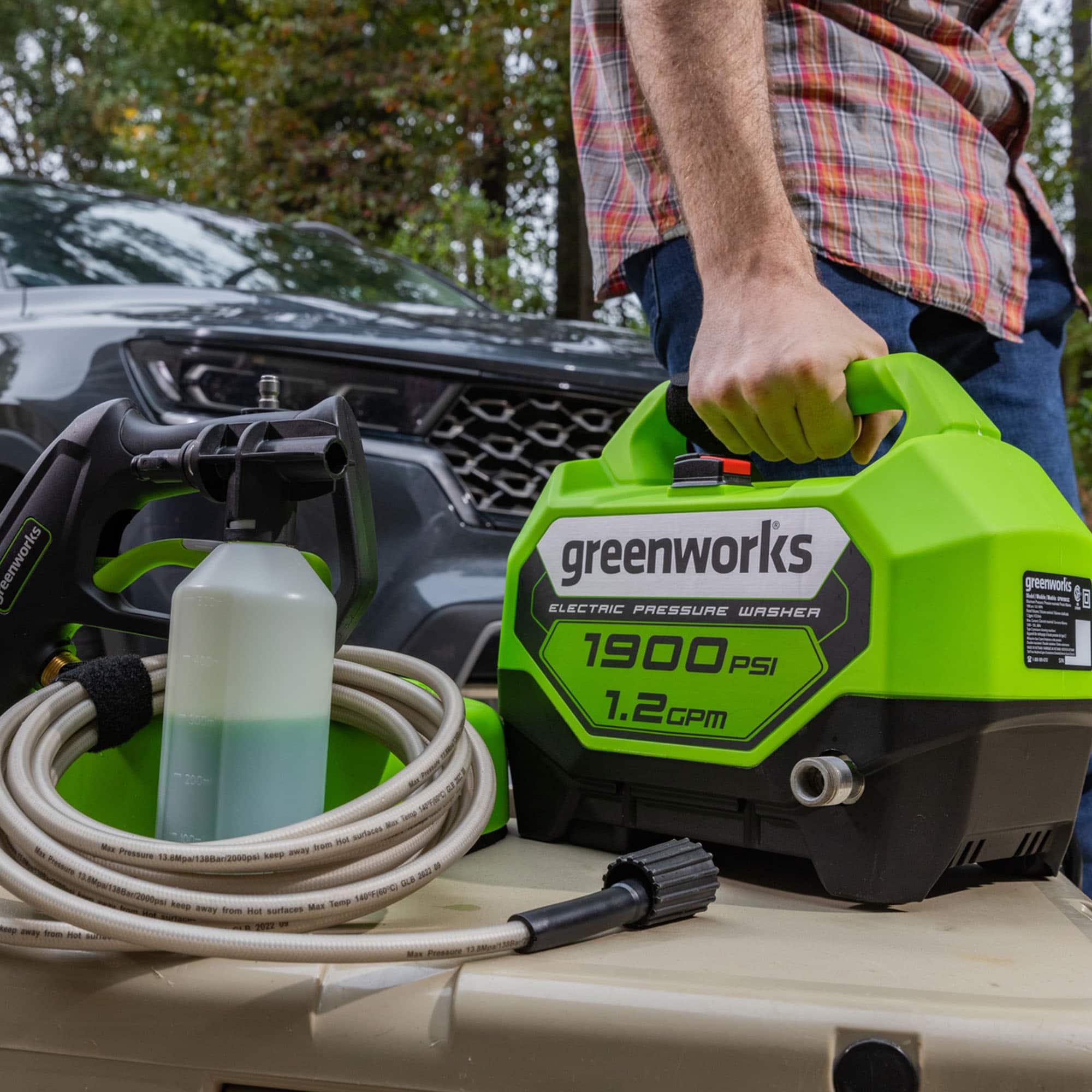 A man is using a Greenworks electric pressure washer to clean his car. The pressure washer is connected to a hose and is being held by the man. The car is parked nearby, and the scene suggests that the man is taking care of his vehicle by using the pressure washer to remove dirt and debris.