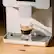 The image features a small coffee maker with a cup of coffee on top of it. The coffee maker is white and has a digital display. The cup of coffee is placed on a wooden table, and the coffee maker is sitting on the table as well. The coffee maker is designed to make espresso drinks, and it is a compact and convenient appliance for preparing coffee at home.