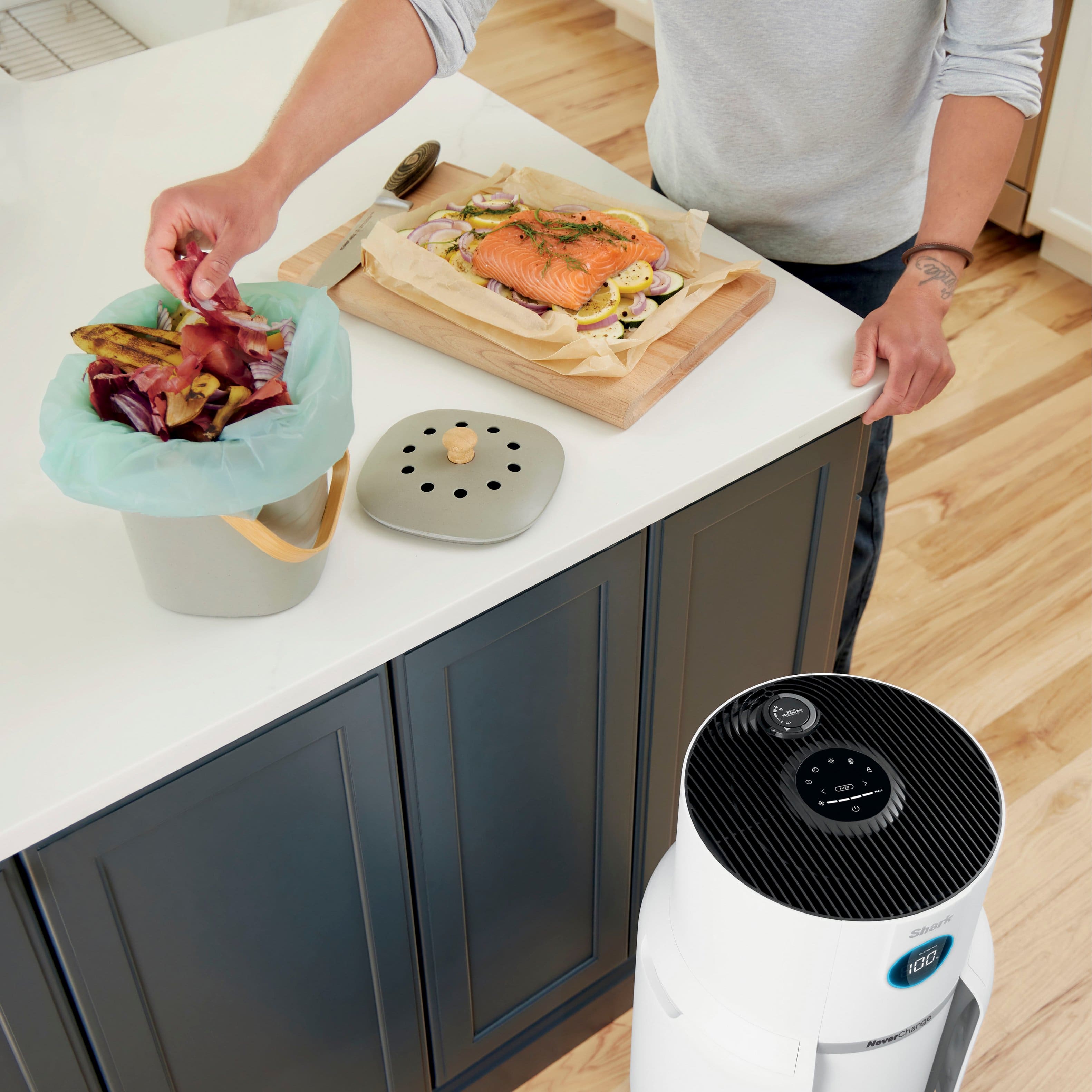 The image features a person standing in a kitchen, preparing food. The person is holding a cutting board with fish on it, and there is a bowl nearby. The kitchen counter has a cutting board, a knife, and a bowl on it. Additionally, there is an oven in the background. The scene suggests that the person is cooking and dealing with lingering cooking odors. The image is promoting an Odor Neutralizer Technology that releases a fresh scent to help combat such odors.