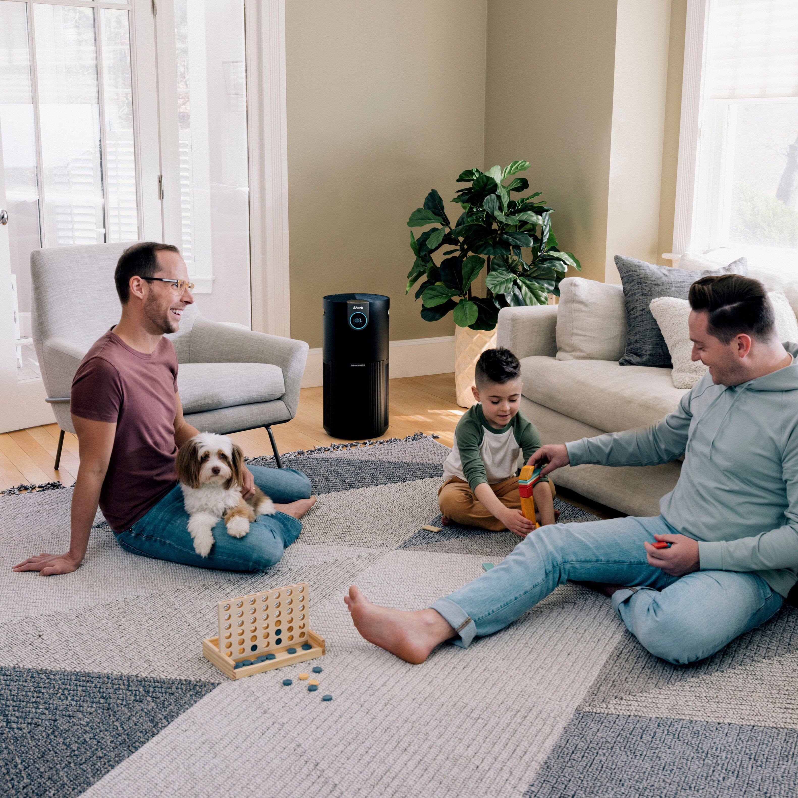 The image shows a family sitting on the floor in a living room, with a dog nearby. There is a potted plant in the room, and a chair is also present. The family appears to be enjoying their time together. The living room is covered with a 1200 sq. ft. of coverage, making it perfect for living rooms, dining rooms, and basements.