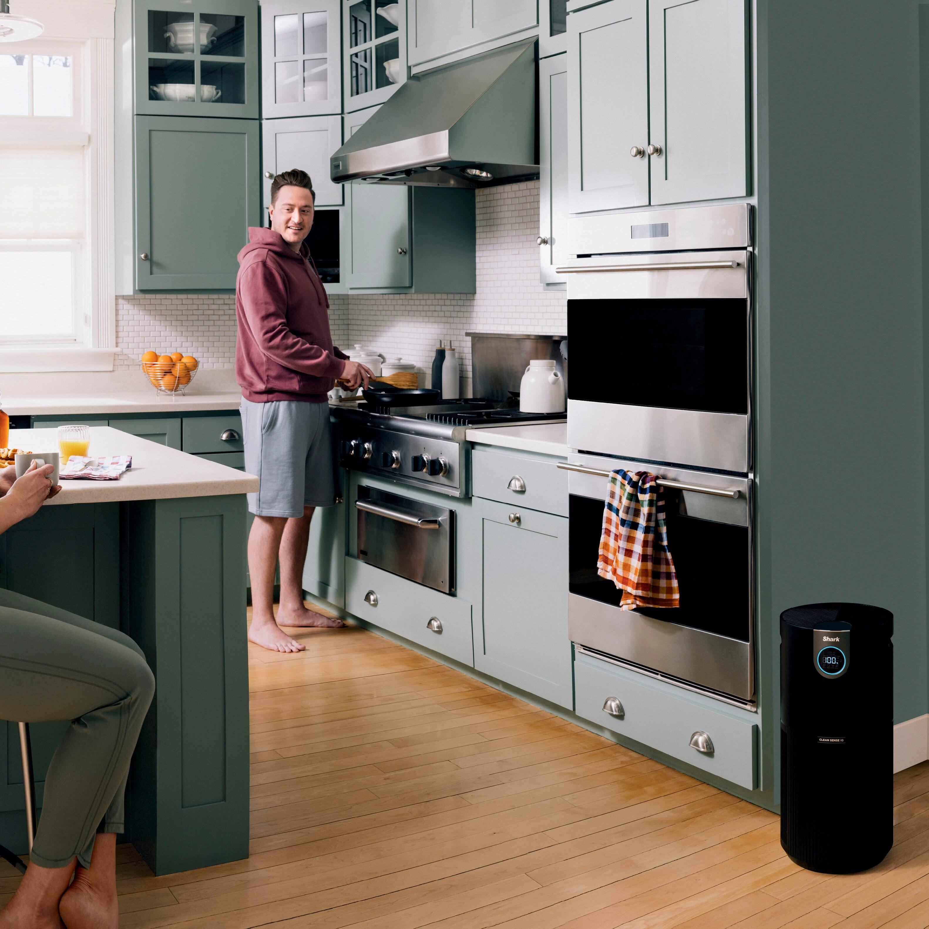 The image depicts a man and a woman in a kitchen, with the man standing near the stove and the woman sitting at the counter. The kitchen is equipped with a stove, oven, sink, and various items such as bowls, cups, and oranges. The man is wearing a red hoodie, and the woman is wearing a white shirt. The scene suggests a cozy and comfortable atmosphere as the couple enjoys their time together in the kitchen.