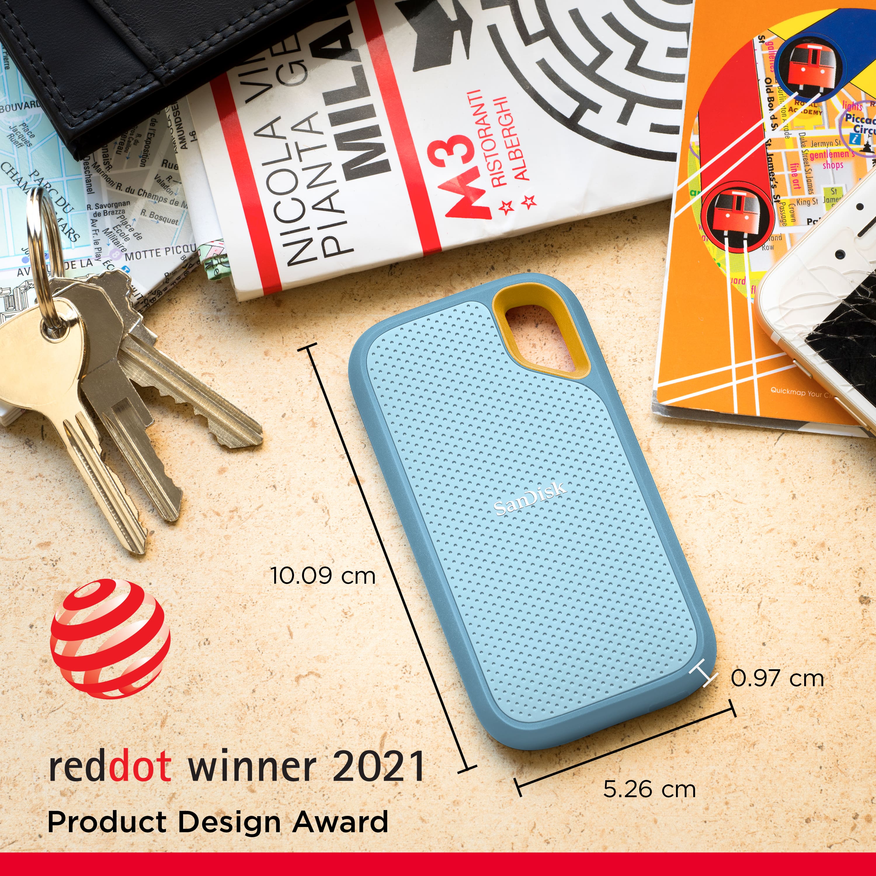 The image features a blue cell phone case with a keychain attached to it. The case is placed on a table, and there are several books and a wallet nearby. The cell phone case is described as a product design award winner, and its dimensions are given as 10.09 cm and 0.97 cm. The image also includes a keychain, which is attached to the cell phone case.