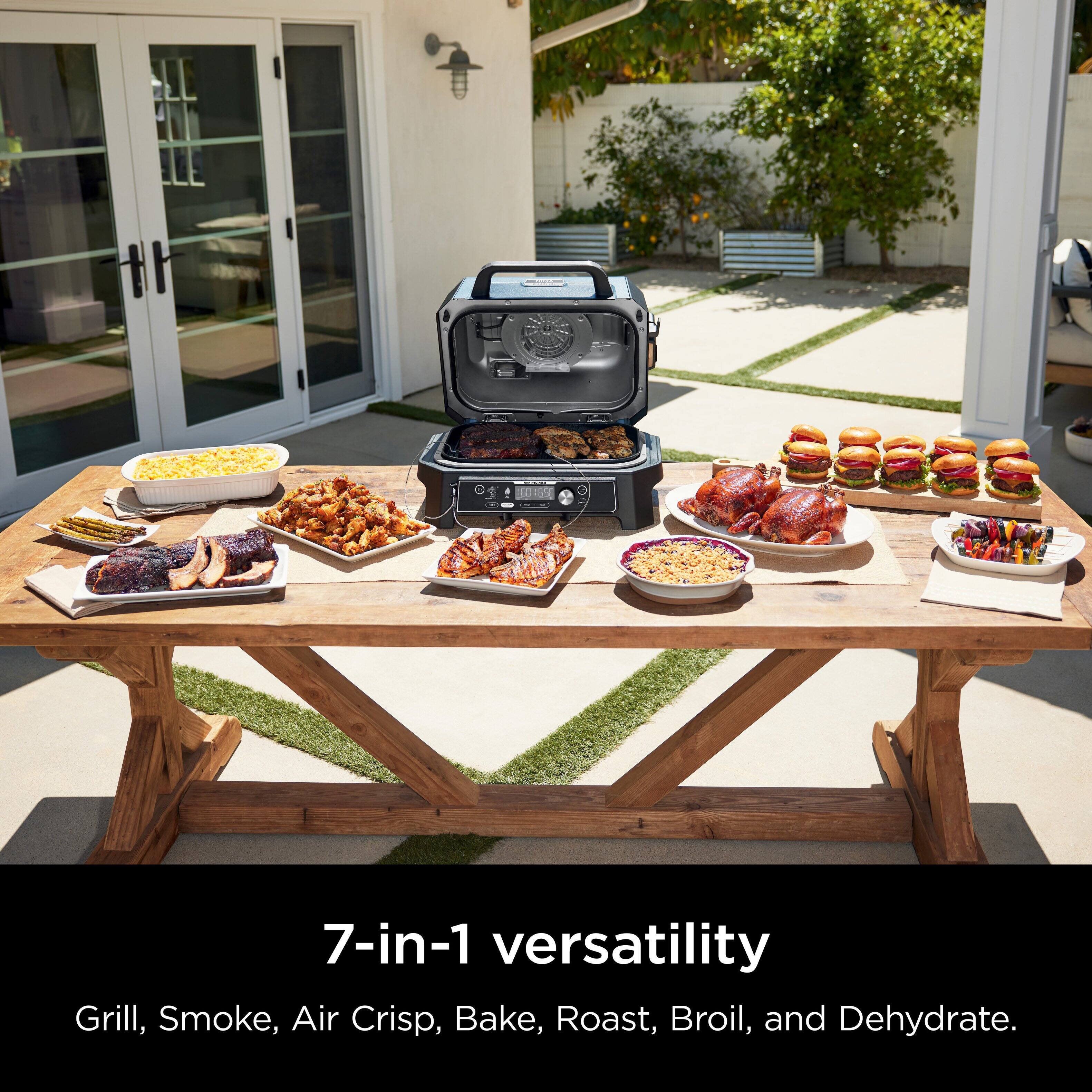 The image features a table filled with various food items, including grilled chicken, corn on the cob, and other dishes. The table is set up for a barbecue, and there is a 7-in-1 versatility grill on display, which can be used for grilling, smoking, air crisping, baking, roasting, broiling, and dehydrating. The table is surrounded by chairs, and there is a potted plant nearby, adding to the outdoor ambiance.