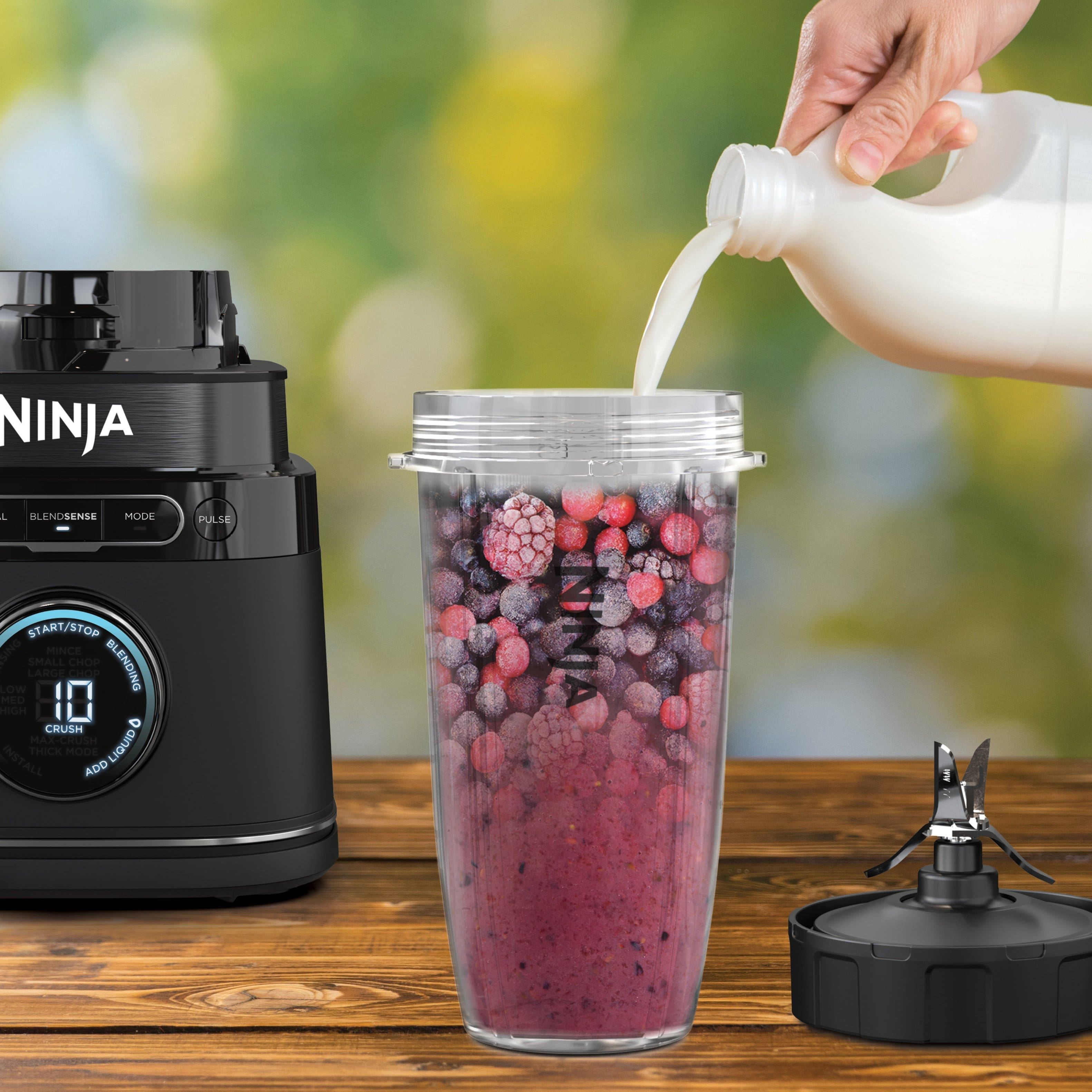 The image features a Ninja blender with a cup of berries inside, and a bottle of milk being poured into the blender. The blender is placed on a wooden table. The text "Liquid Detect Alerts you when liquid is needed so you always get silky-smooth drinks" is displayed in the foreground, likely promoting the blender's features and benefits.
