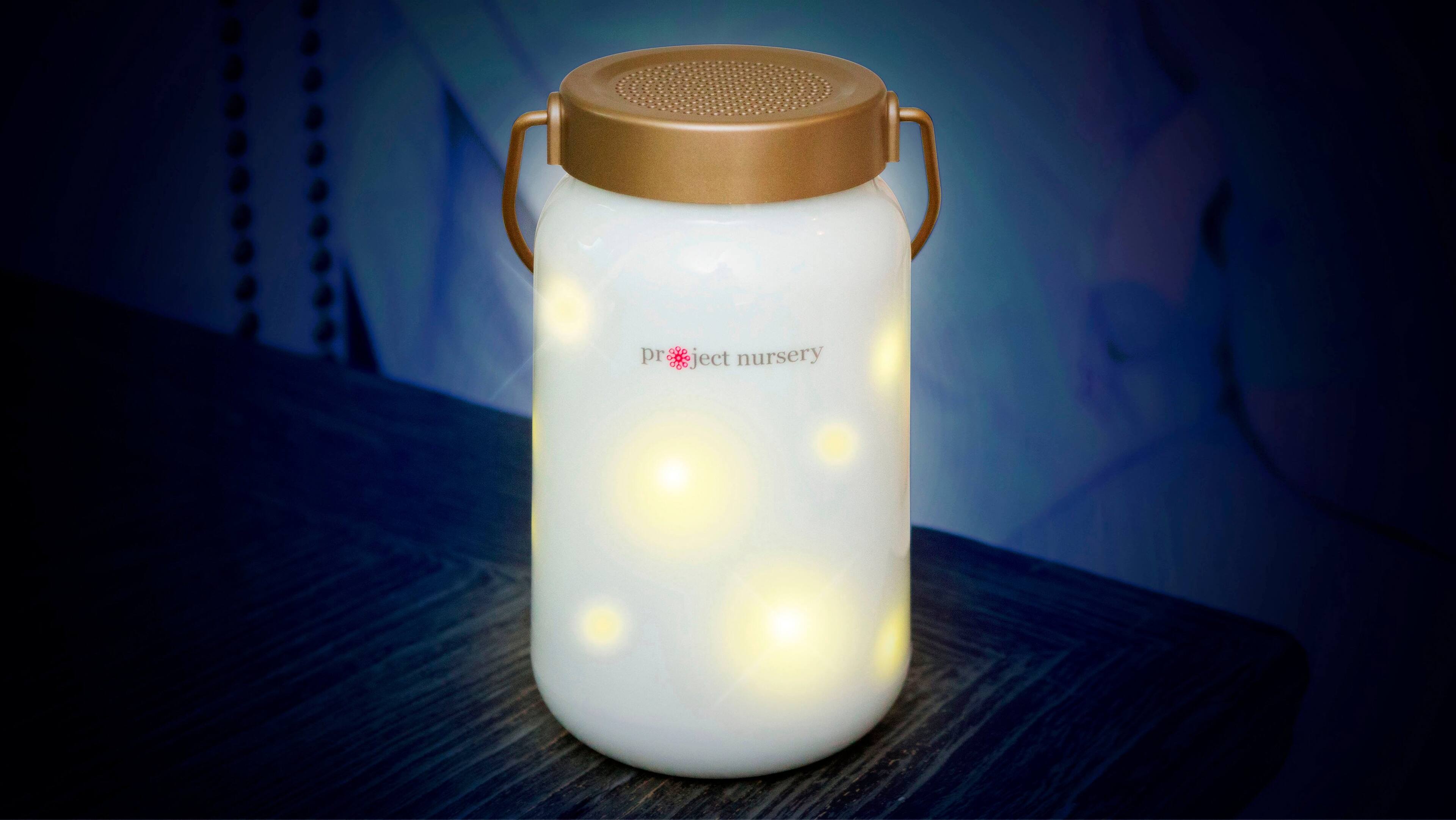 The image features a white jar with a gold lid, which is filled with a white substance and has a light inside it. The jar is placed on a wooden table, and it appears to be a soother or a sound machine. The light inside the jar is likely used to create a calming atmosphere for babies or to help people sleep. The jar is labeled with the text "pr ject nursery," which suggests that it is a product designed for use in a nursery or a child's bedroom.
