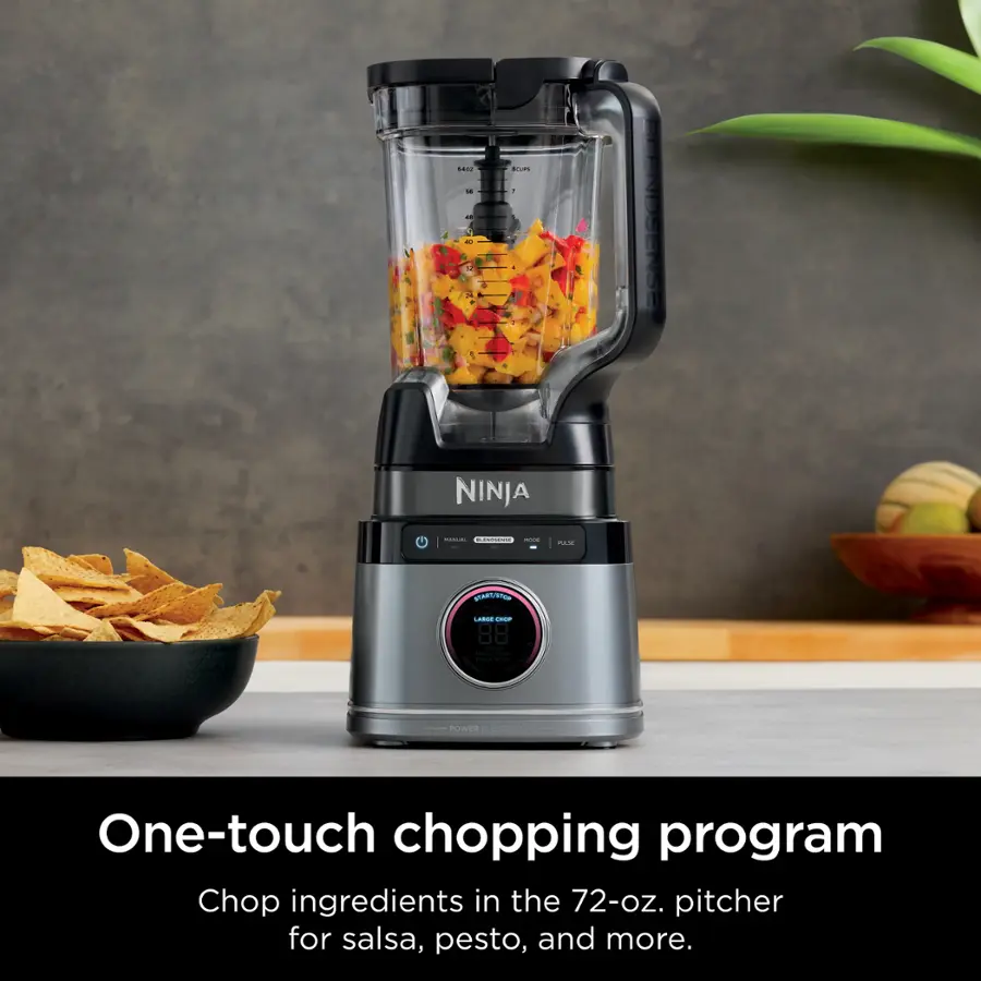 The image features a Ninja chopper, which is a one-touch chopping program that chops ingredients in the 72-oz. pitcher for salsa, pesto, and more. The chopper is placed on a counter, and there is a bowl of chips nearby. The scene is set against a backdrop of a potted plant, adding a touch of greenery to the space.
