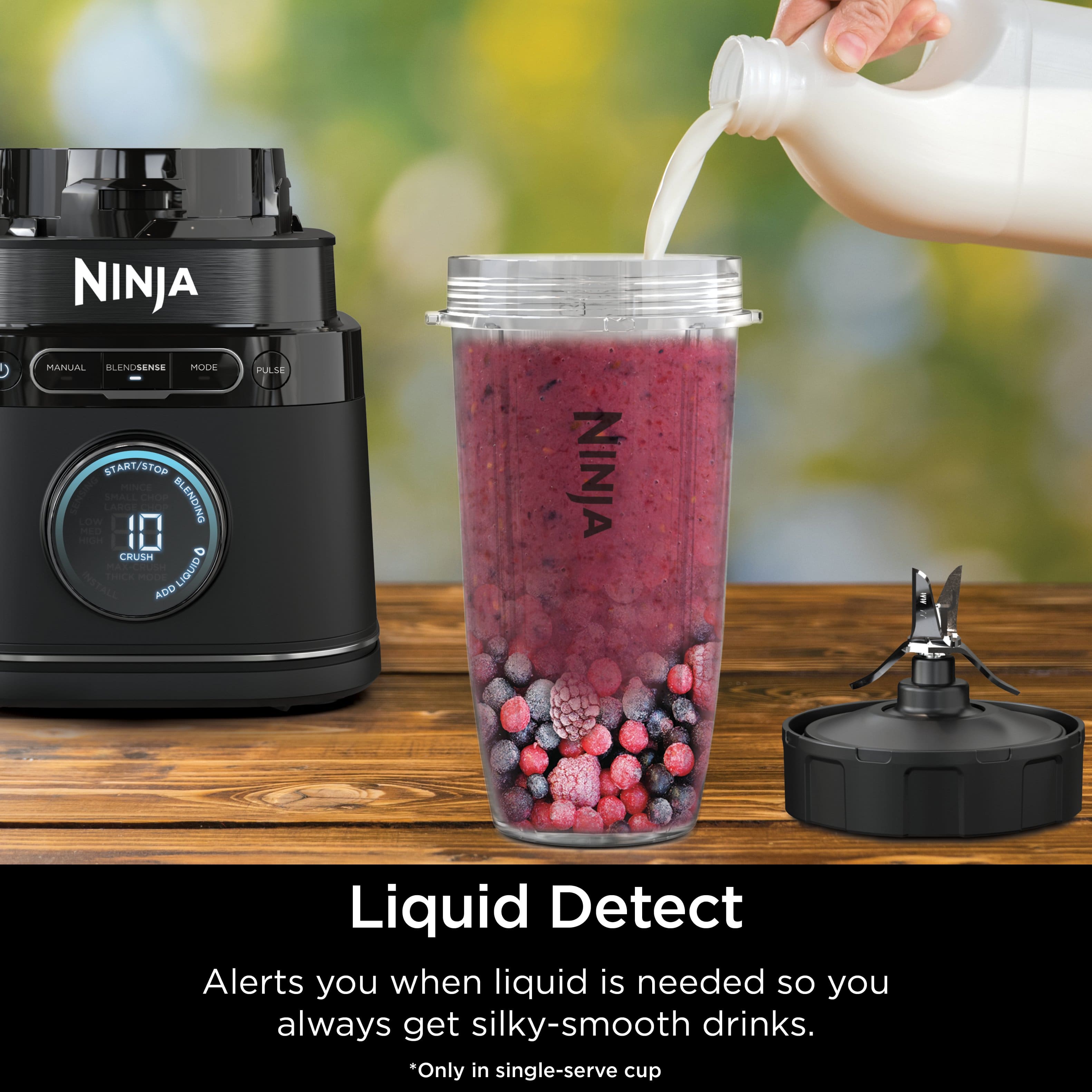 The image features a Ninja blender with a container of liquid being poured into it. The blender is filled with blueberries and raspberries, indicating that a smoothie is being prepared. The blender is placed on a wooden table, and the Ninja manual is also visible nearby. The image highlights the Liquid Detect feature of the Ninja blender, which alerts users when liquid is needed to achieve a silky-smooth drink. This feature is only available in single-serve cups.