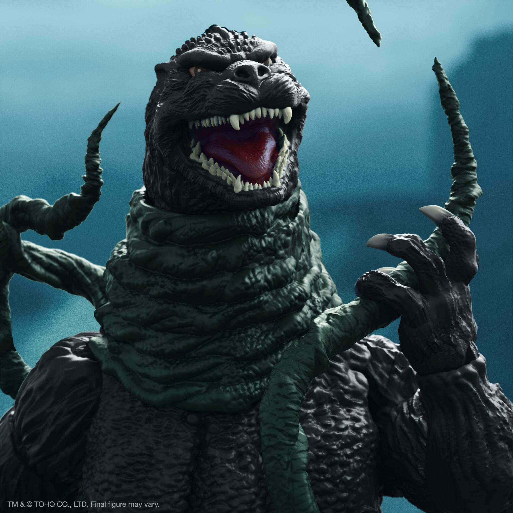 The image features a large action figure of a monster, likely from the Godzilla franchise, with its mouth open and teeth showing. The figure is wearing a green toga and has its arms outstretched. The monster's face is prominently displayed, with its teeth and tongue visible. The final figure may vary, as it is a product of TM & TOHO CO., LTD.
