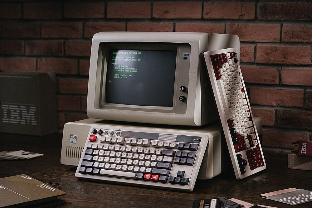 The image shows an old-fashioned IBM computer with a keyboard and a monitor on a desk. The computer is set up next to a brick wall, and there is a book nearby. The text on the image reads: "a3 #--205 Ctc XCr Reclle 1M 0ggtight for Drp IE Jctbdri I ent fllm 16 bycm notal dint CH cotin ena a byt Botal ary S4Z2 hyte Ime a IBM 2 : DM IBM y 0 A Z a W 8 X 2 a E D a - A R F V - T e B - Y H N " , U J M P a / K . O E a 2 - - P 7 P " 1 1 P - ~ 0 1 IBM IBM B A A."