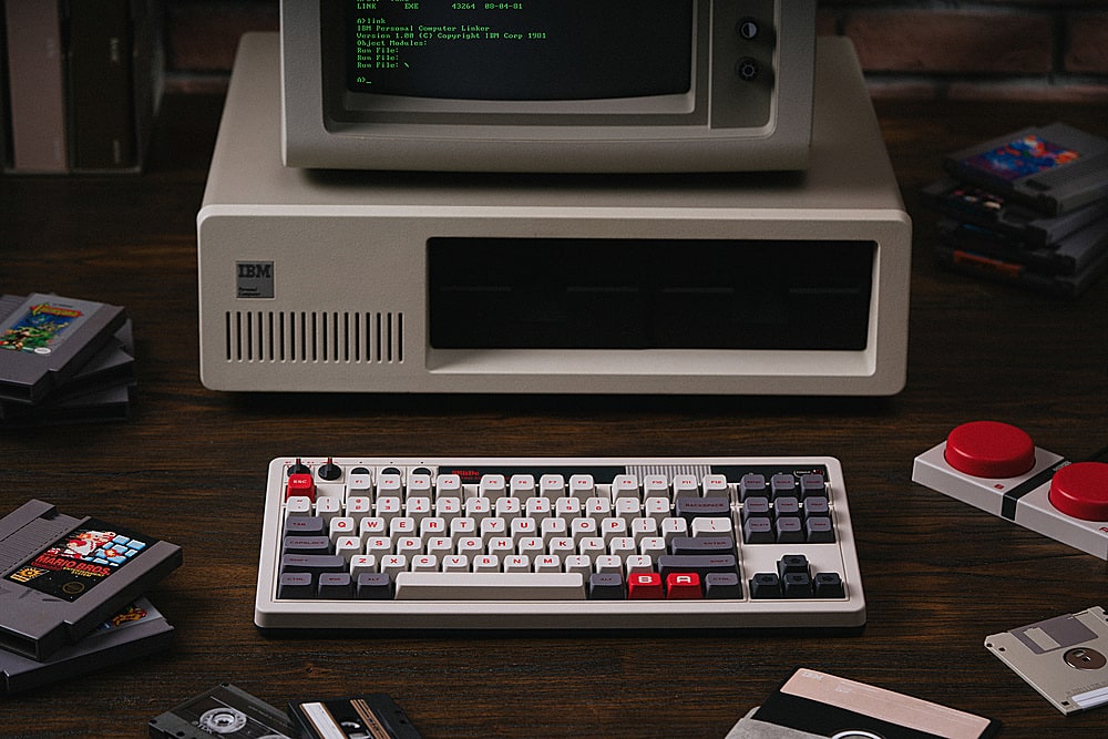 The image features a vintage computer setup, including a keyboard, a monitor, and a collection of Nintendo game cartridges. The keyboard is placed in front of the monitor, and the game cartridges are scattered around the area. The monitor is displaying text, which reads "LIKE EXE 43244 10-8-0 limh 1tmm Personal Computor Linker Verste 1.00 (C) Copyright 1901 Ohject Modules Eun Fille: Eun Filo: Kum File: I A>. IBM ... 1 - - E - - - - a . - a 7 . - A 1 . + - . Q W E F T Y U I O 1 1s H P4 J K T L - ! 1 - - A."