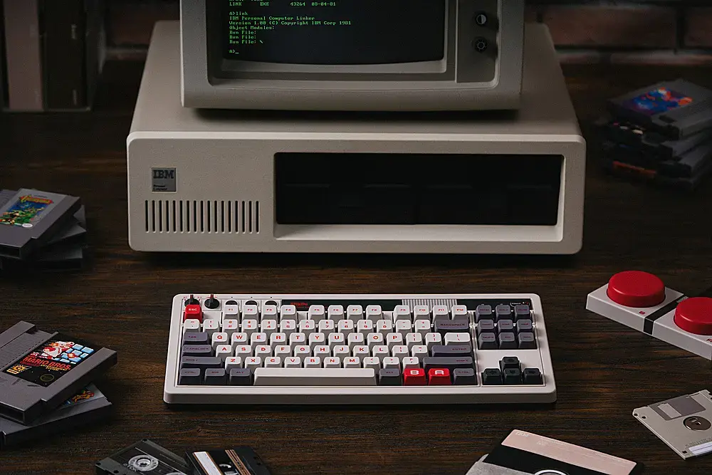 The image features a vintage computer setup, including a keyboard, a monitor, and a collection of Nintendo game cartridges. The keyboard is placed in front of the monitor, and the game cartridges are scattered around the area. The monitor is displaying text, which reads "LIKE EXE 43244 10-8-0 limh 1tmm Personal Computor Linker Verste 1.00 (C) Copyright 1901 Ohject Modules Eun Fille: Eun Filo: Kum File: I A>. IBM ... 1 - - E - - - - a . - a 7 . - A 1 . + - . Q W E F T Y U I O 1 1s H P4 J  K T L - ! 1 - - A."