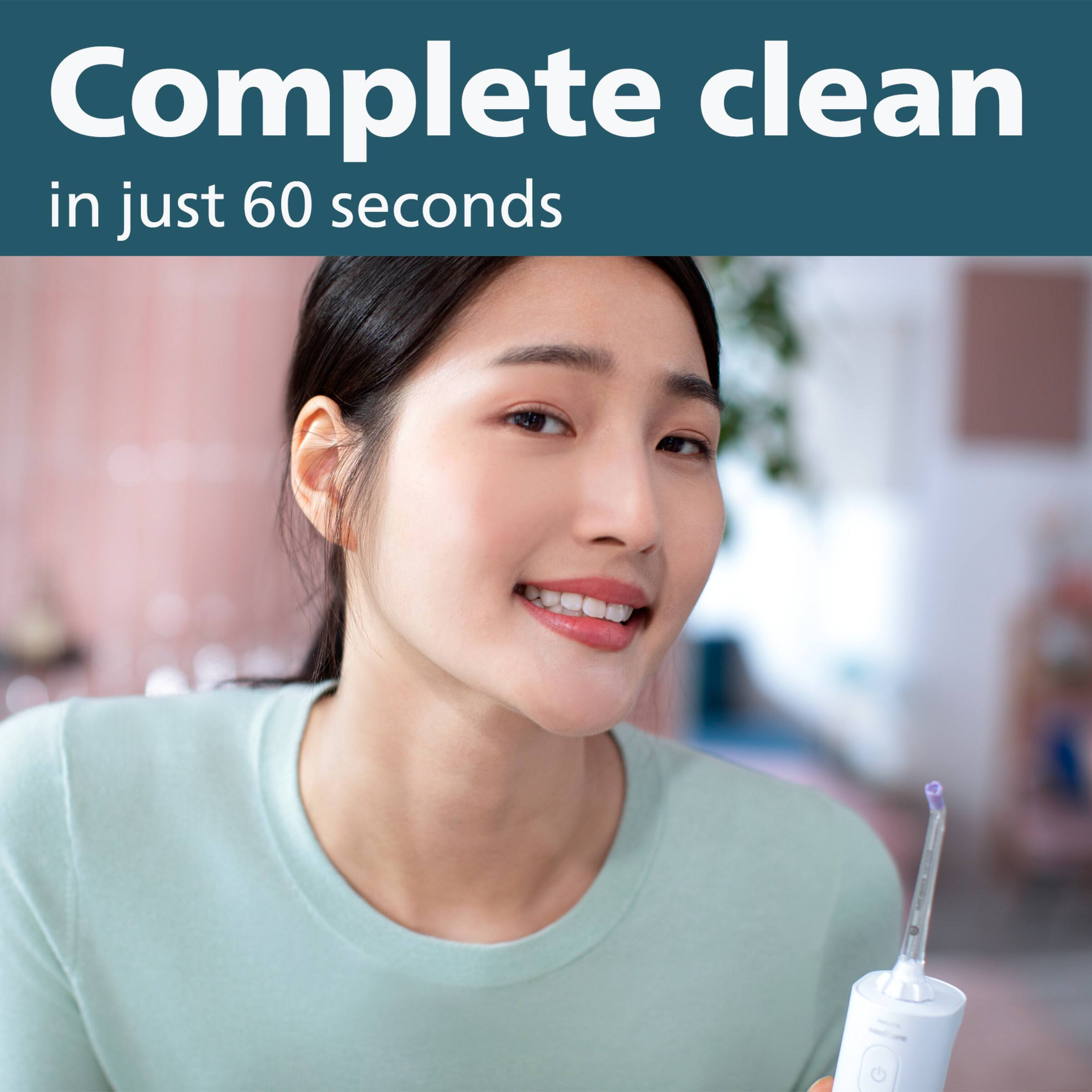 The image features a woman smiling and holding a water flosser in her mouth. The text "Complete clean in just 60 seconds" is displayed above her head, emphasizing the efficiency of the water flosser. The woman is wearing a green shirt, and there is a potted plant in the background.