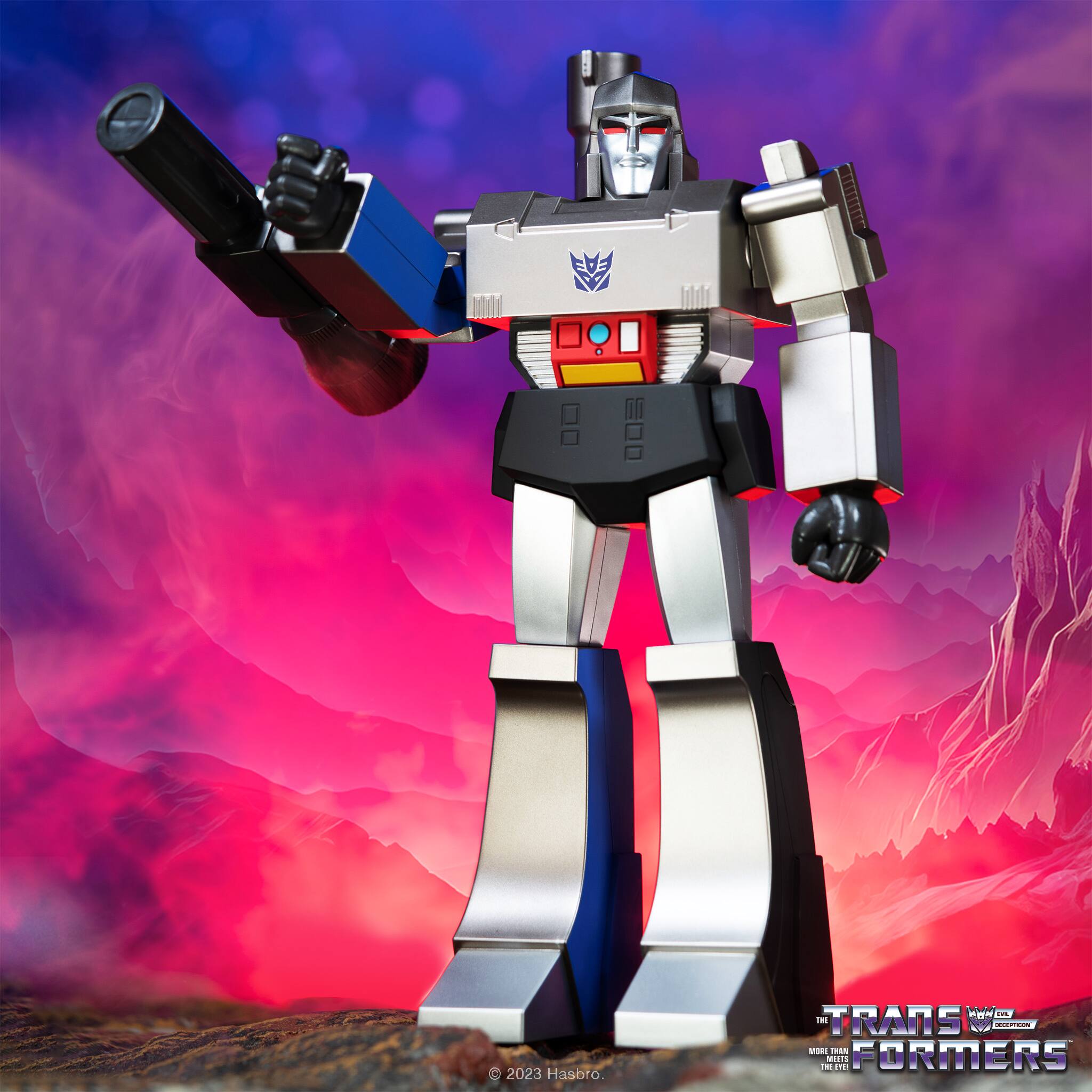 The image features a robotic figure, likely an action figure, standing in a red and purple background. The figure is holding a gun, and it appears to be a Transformers toy. The toy is positioned in a way that it seems to be looking at the camera. The background consists of a mountainous landscape, adding to the overall visual appeal of the scene.