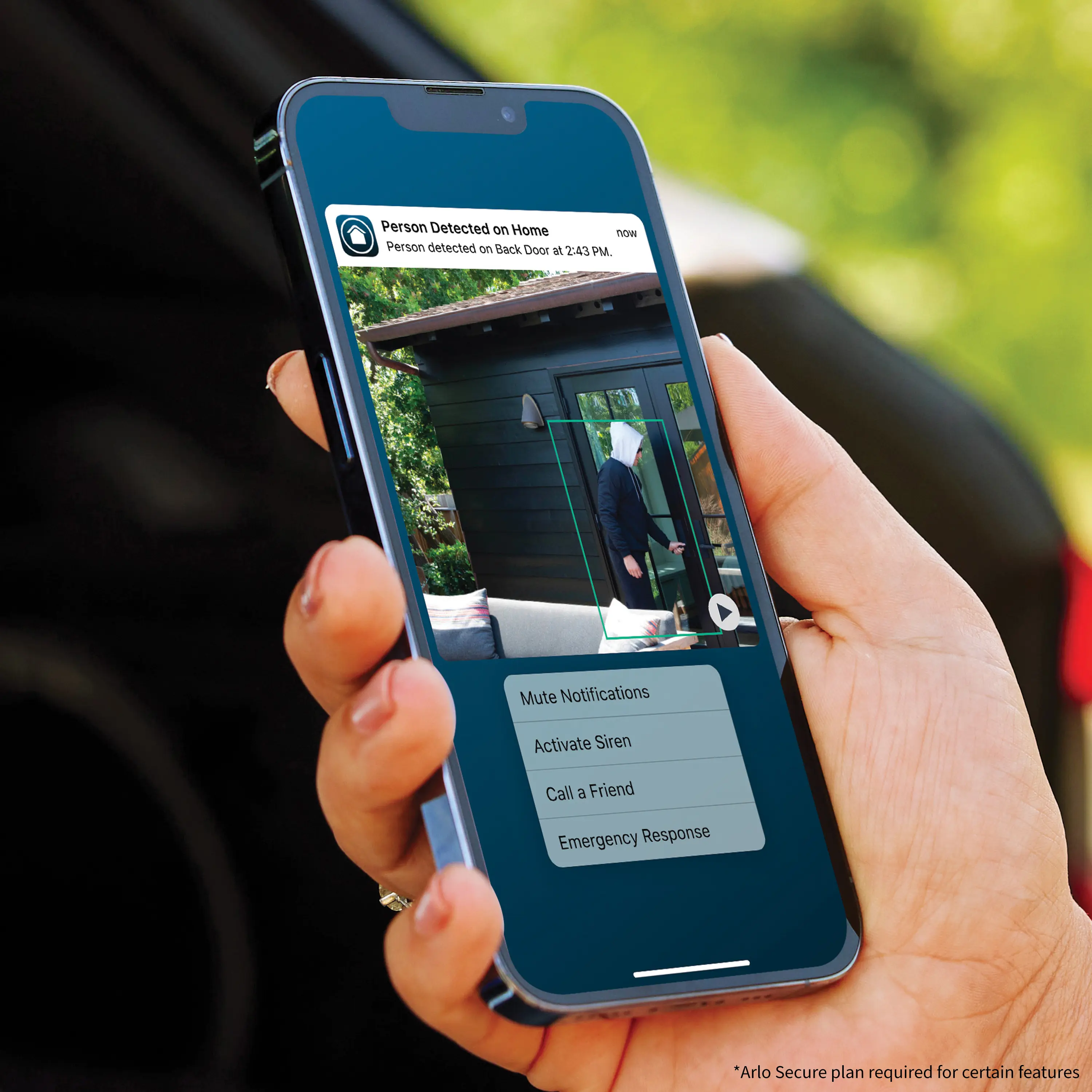 The image shows a person holding a cell phone with a live video feed displayed on the screen. The video feed shows a person standing in front of a house. The cell phone is being used to monitor the situation, possibly for security purposes. The person holding the phone is likely using an app or service that allows them to view the live video feed from a remote location.