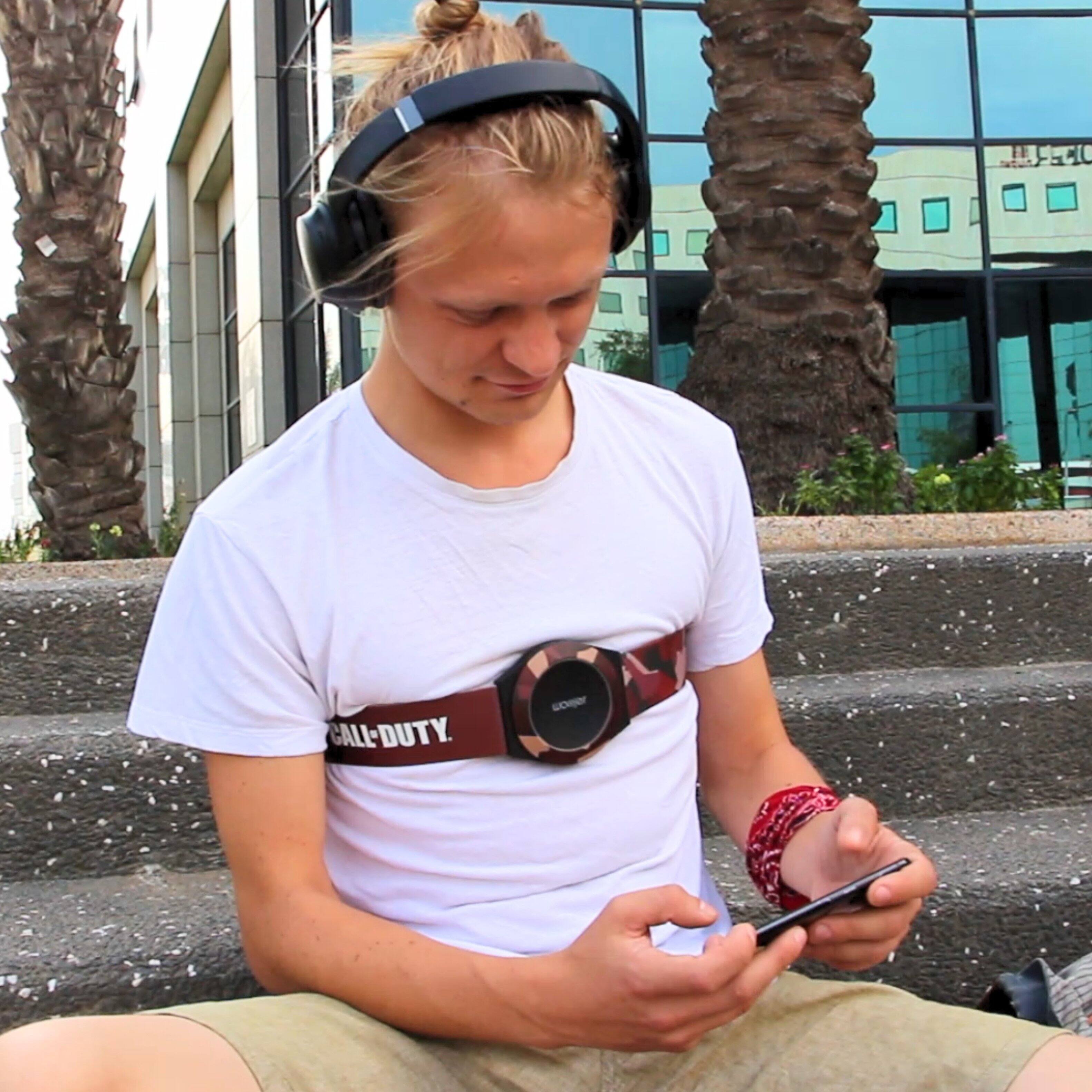 The man is wearing a Call of Duty belt buckle and headphones. He is sitting on a bench and looking at his phone.
