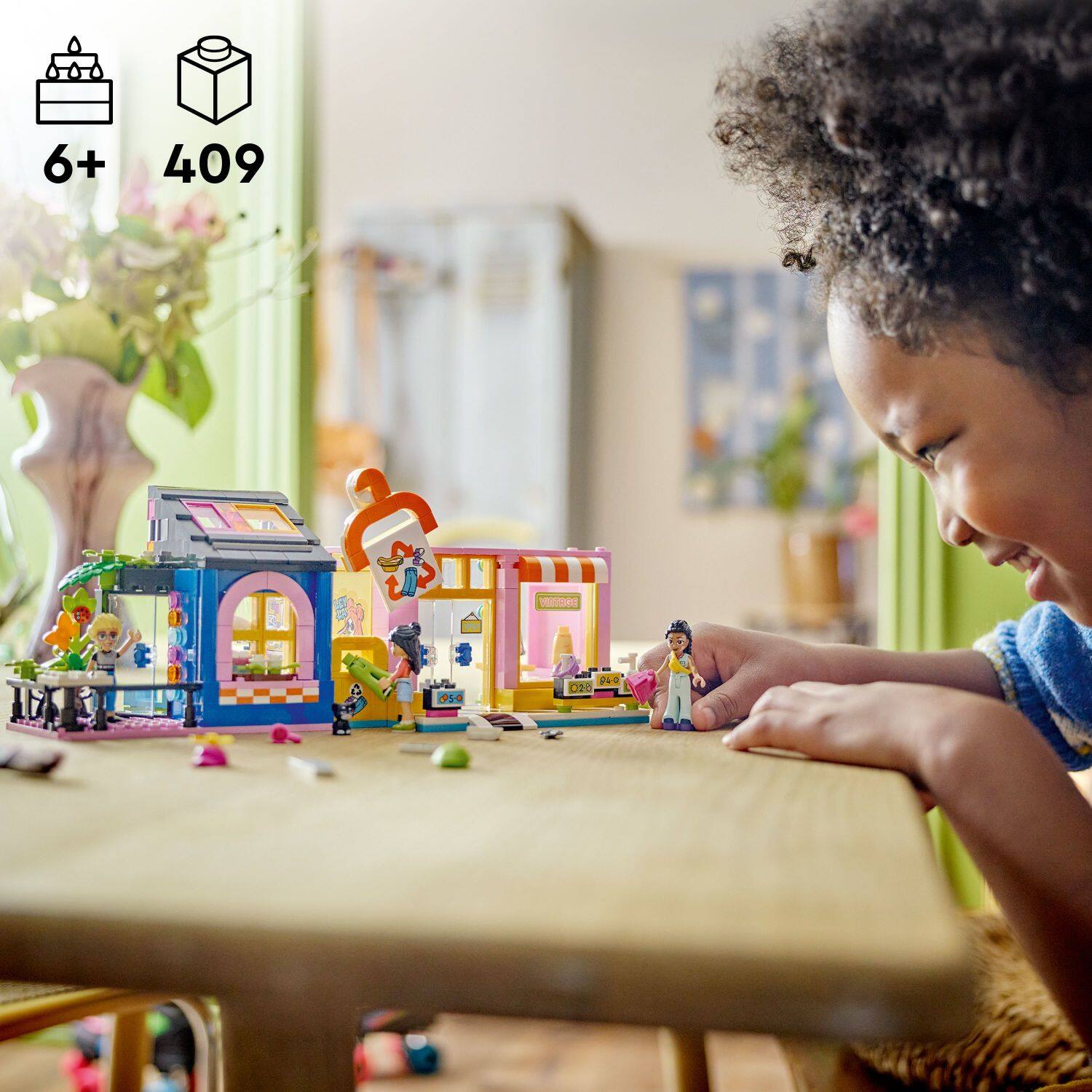 The image features a young girl playing with a toy set, which includes a toy house and a toy car. She is sitting at a table and appears to be enjoying herself while engaging with the toys. The toy set appears to be a building set, as it includes various components for constructing and creating different structures. The girl's smile and focused attention on the toys suggest that she is having a fun and creative playtime.