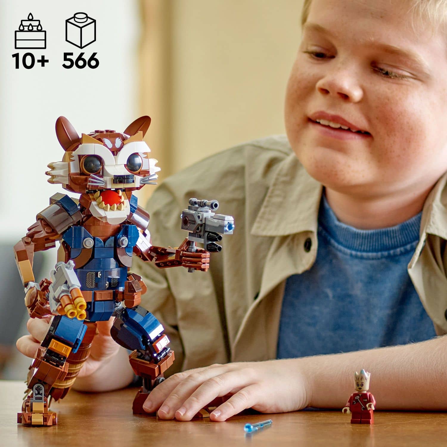 The image features a young boy holding a Lego figure, possibly a robot or a character from a popular franchise. The boy is smiling and appears to be enjoying his time with the toy. The Lego figure is positioned in front of the boy, and he is holding it with both hands. The scene takes place on a dining table, which occupies most of the background.