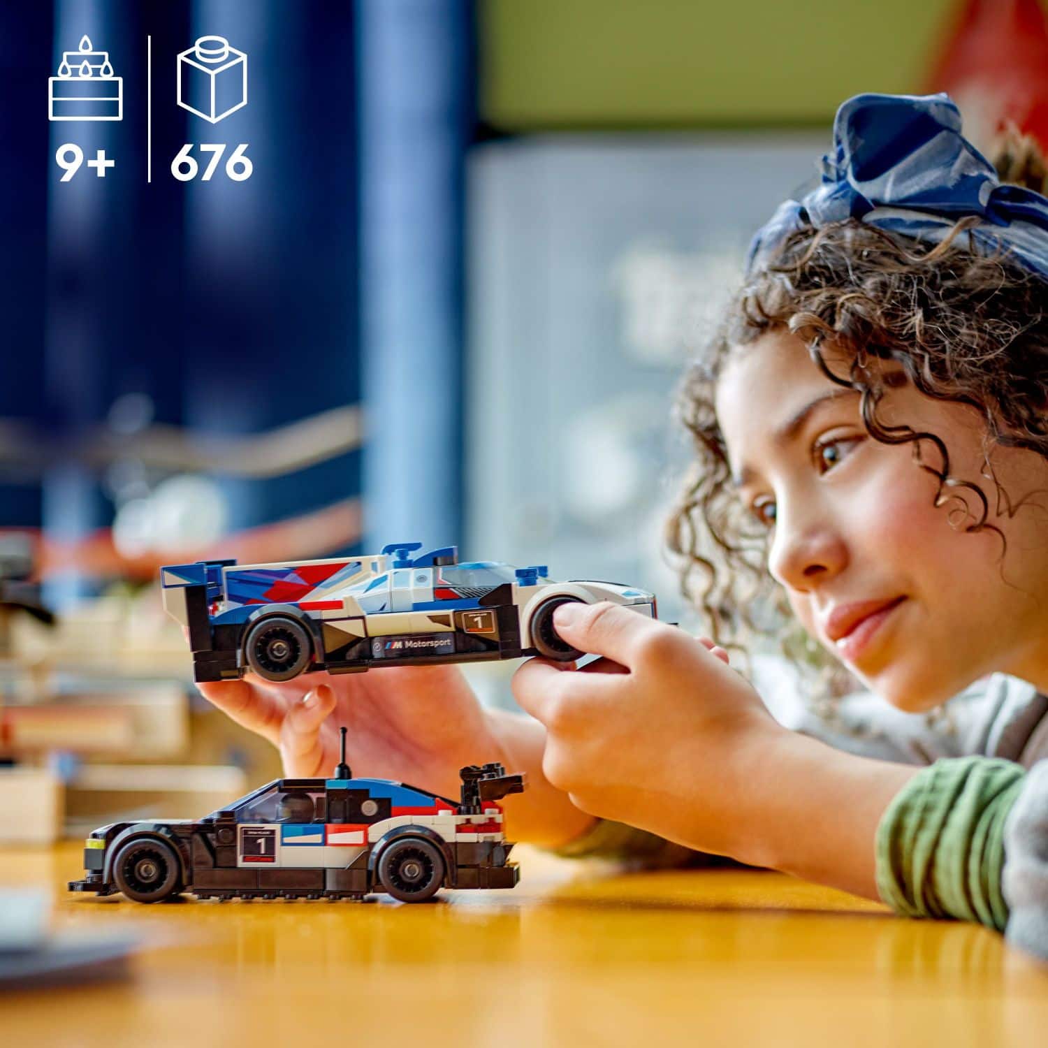 The image shows a young girl playing with toy cars, specifically a blue and white race car, on a table. She is holding the toy car and appears to be enjoying her time with the toy. The scene is categorized as Blocks and Building Sets, which suggests that the toy cars might be part of a building set or a construction toy.