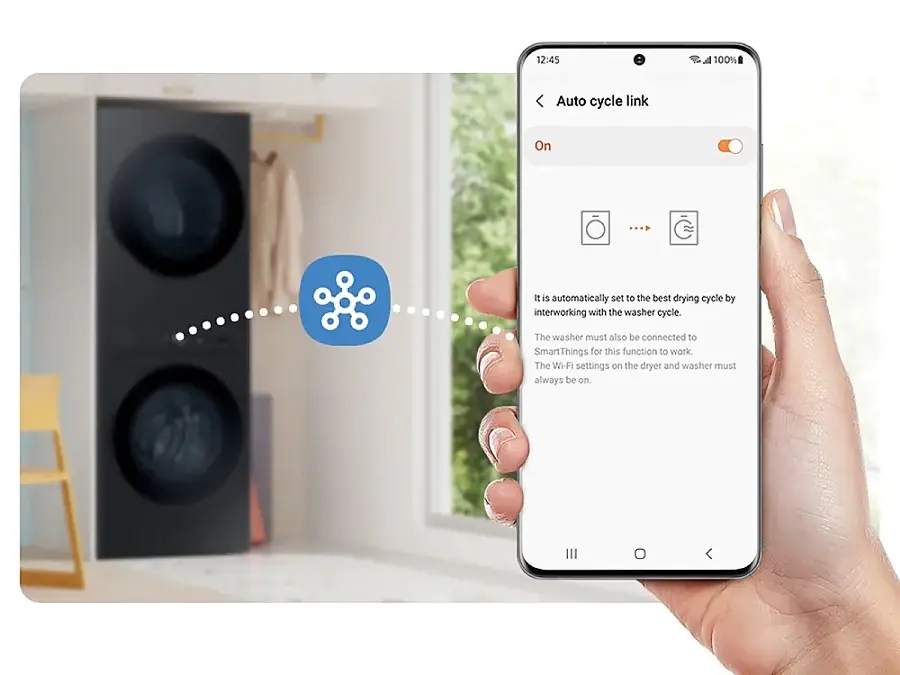 The image shows a person holding a cell phone with a message displayed on the screen. The message reads: "12:45 a 100% Auto cycle link On ... It is automatically set to the best drying cycle by interworking with the washer cycle. The washer must also be connected to SmartThin Things for this function to work. The Wi-Fi settings on the dryer and washer must always be on."