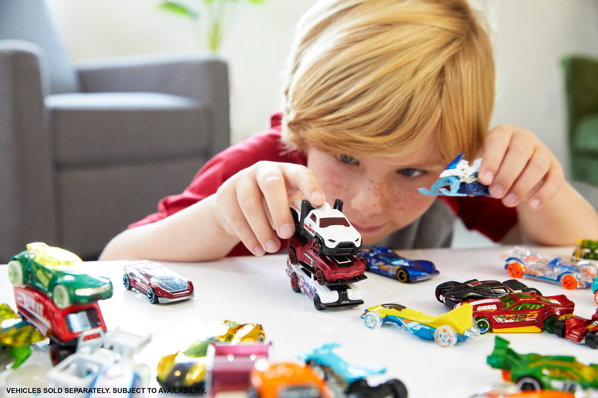 The image shows a young boy playing with toy cars on a table. The toy cars are arranged in a row, and the boy is holding one of them. The text "SXI1 VEHICLES SOLD SEPARATELY. SUBJECT TO AVAILABILITY." is not relevant to the scene and can be removed.