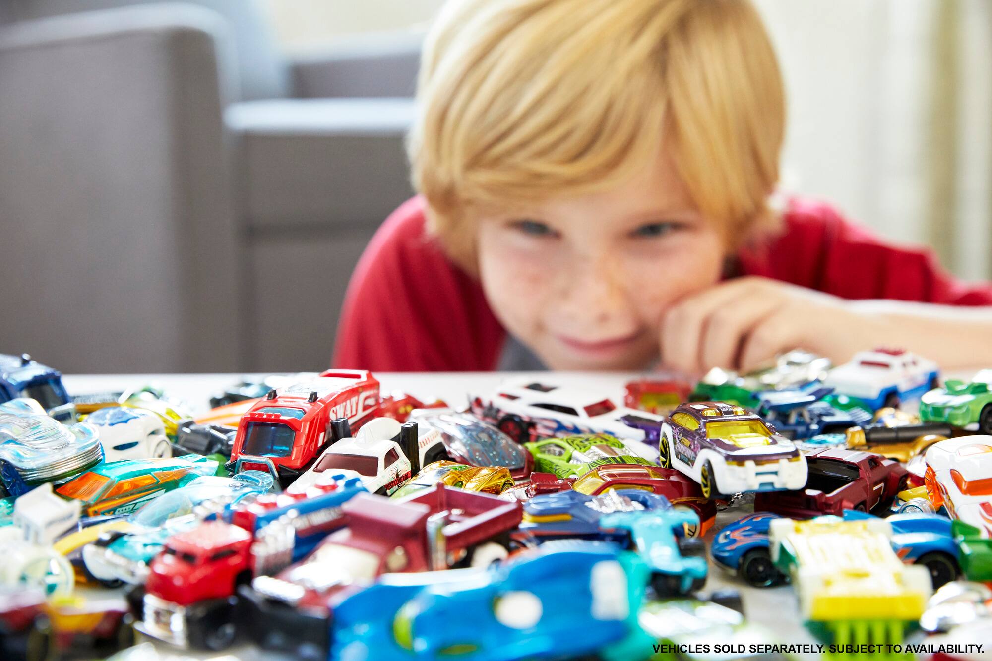 The image shows a young boy sitting in front of a table filled with various toy cars. The toy cars are of different colors and sizes, and the boy appears to be playing with them. The table is covered with numerous toy cars, creating a vibrant and engaging scene. The boy is likely enjoying his time playing with these toy cars, which are available for purchase separately.