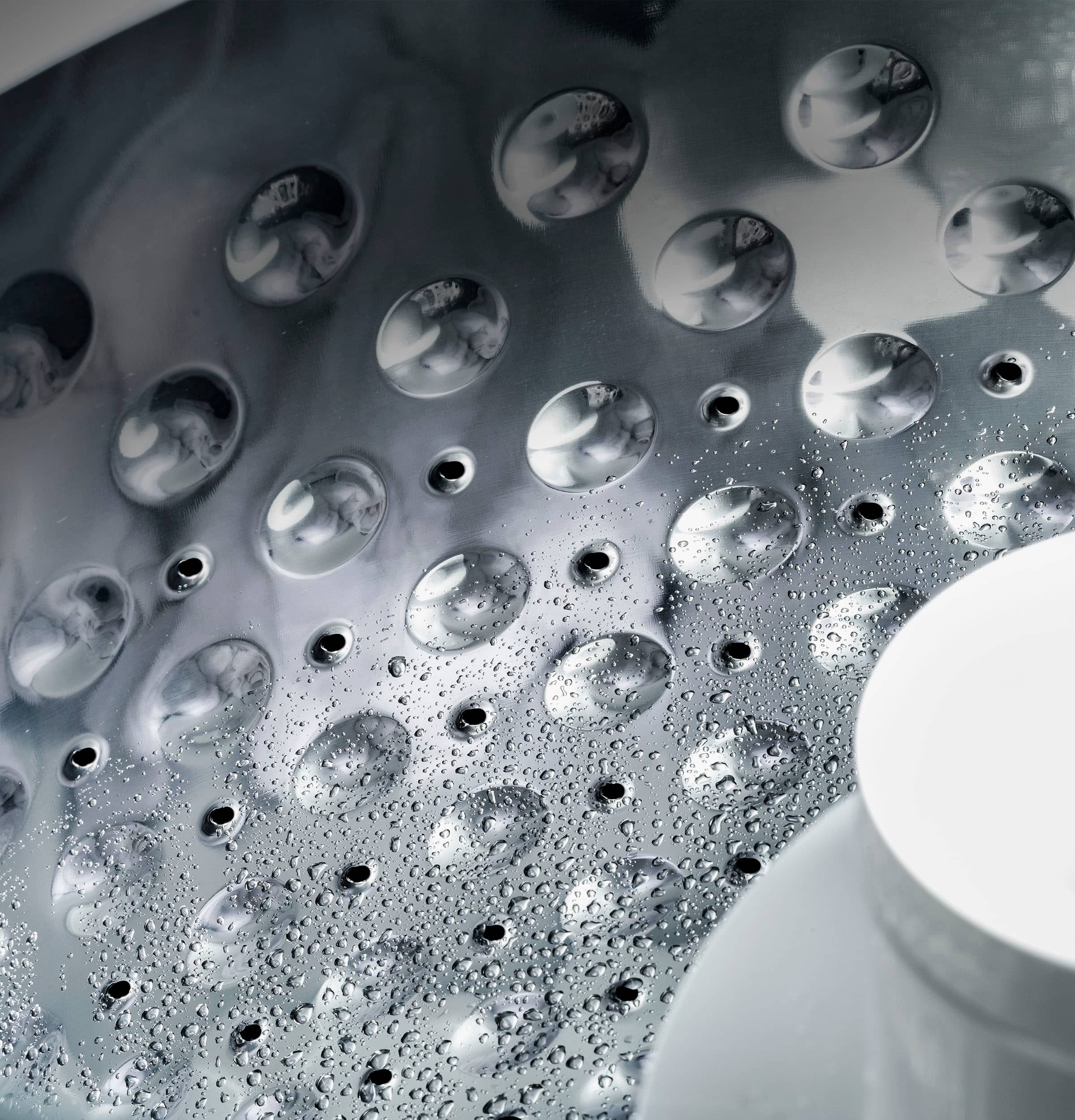 The image features a silver shower head with a pattern of holes on it. The shower head is positioned above a sink, and it appears to be covered in water droplets. The droplets are scattered across the shower head, giving it a shiny appearance. The sink is located to the right of the shower head, and the overall scene creates an impression of a well-maintained bathroom.