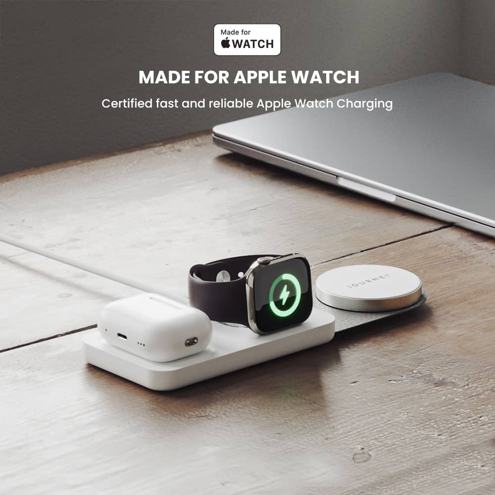 The image features a wooden table with a laptop and various electronic devices. There is a watch charging station with a watch on it, and a cell phone is also present on the table. The scene suggests that the devices are being charged or powered, possibly for use with an Apple Watch. The presence of a laptop and cell phone implies that the user might be working or communicating while charging their devices.