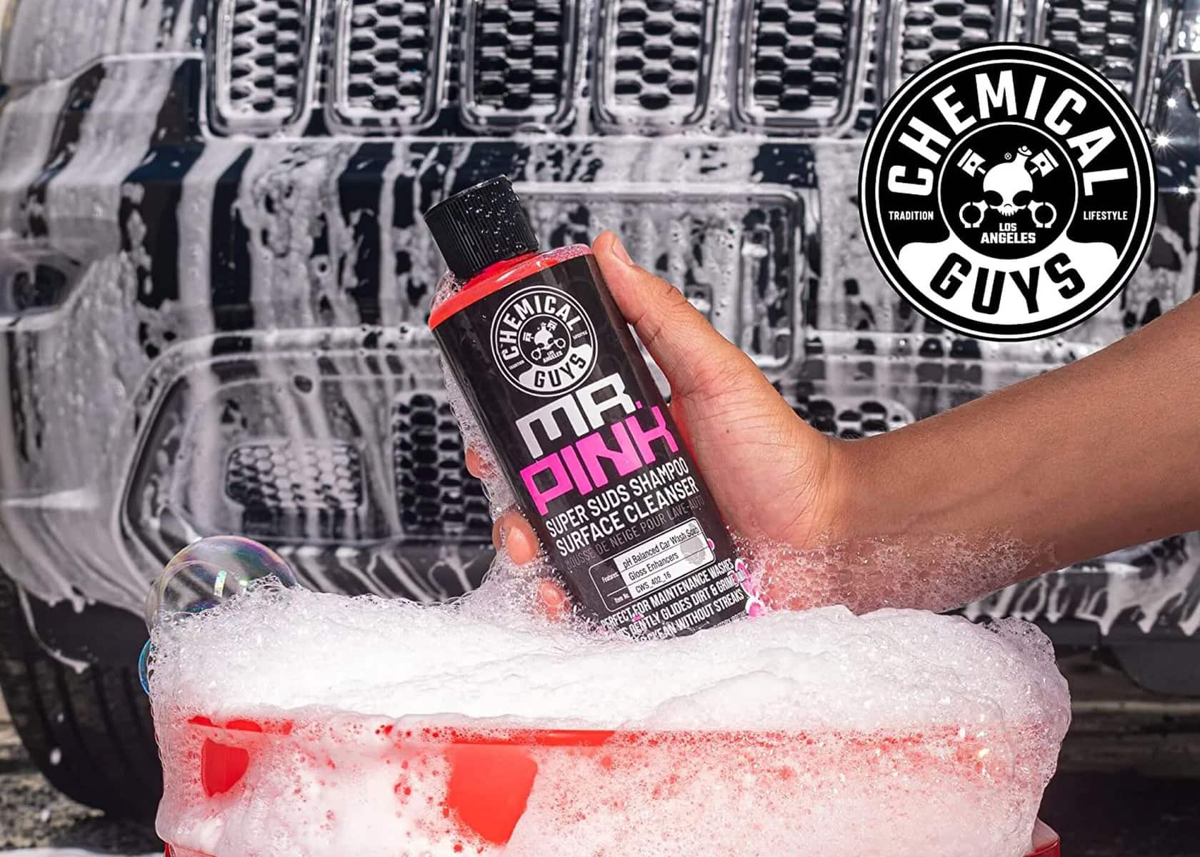 The image features a person holding a bottle of Mr. Pink car wash soap, which is a product from the Chemical Guys brand. The bottle is being used to clean a car, and the suds are being sprayed onto the vehicle. The car being cleaned is a Jeep, and the person is using a spray bottle to apply the car wash solution. The Chemical Guys brand is known for its high-quality cleaning solutions and is popular among car enthusiasts and professionals.