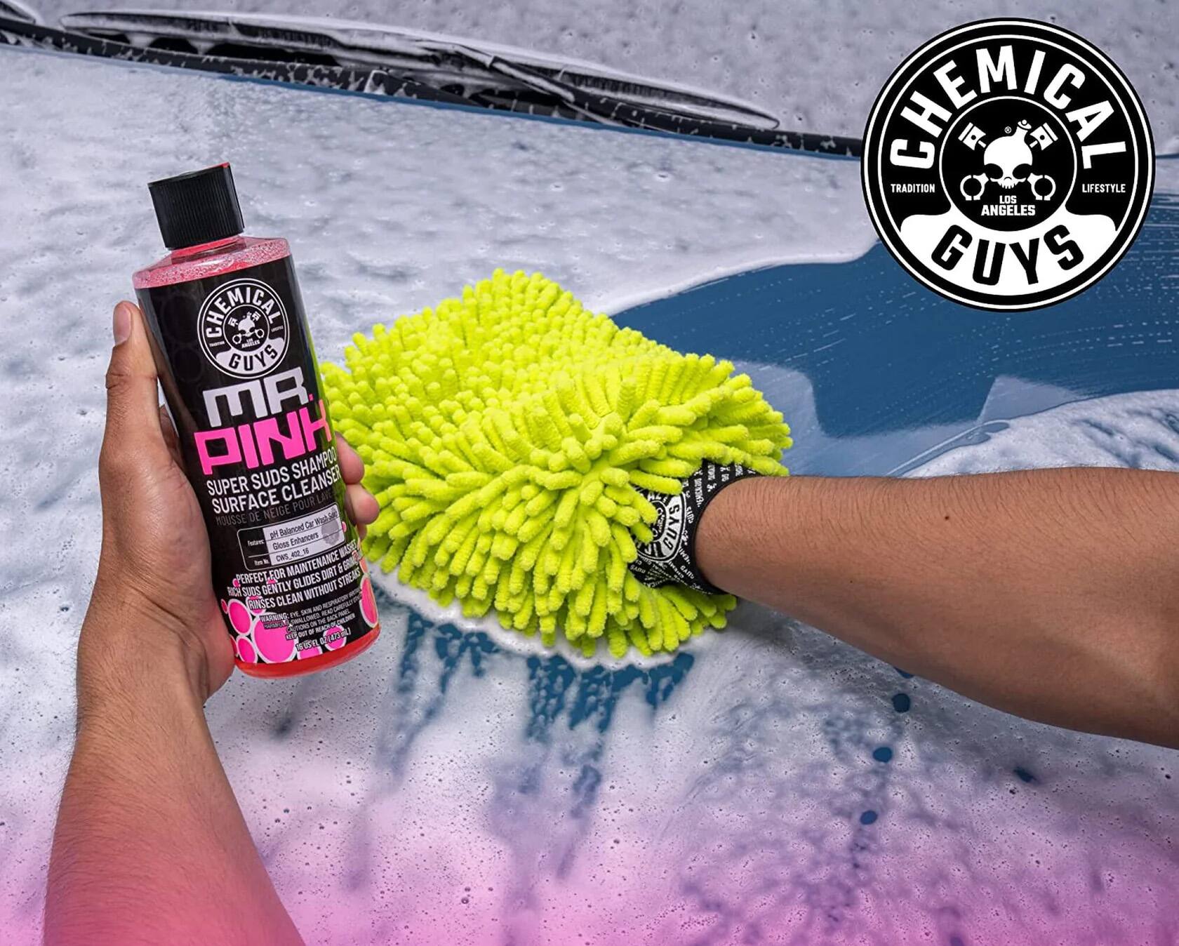 The image features a person holding a bottle of pink cleaning solution, likely a shampoo or surface cleaner, in front of a green scrubbing pad. The person is wearing a watch, and the bottle is positioned close to the scrubbing pad. The scene suggests that the person is using the cleaning solution to clean or maintain the green scrubbing pad. The image is categorized under "Cleaning Solutions" and is likely promoting a cleaning product or showcasing a cleaning technique.