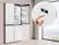 The image shows a kitchen with a refrigerator and a window. The refrigerator is white and has a digital display on the front. A hand is seen pressing a button on the display, possibly adjusting the temperature or selecting a specific setting. The refrigerator is a Samsung model, as indicated by the brand name displayed on the front.