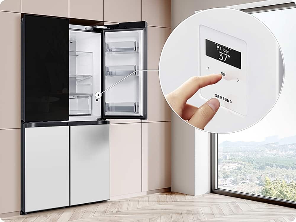 The image shows a kitchen with a large refrigerator and a window. A person is holding a Samsung smart device, which is placed on the refrigerator. The device is likely used to control the refrigerator's temperature or other settings. The refrigerator is empty, and the kitchen appears to be well-maintained and organized.