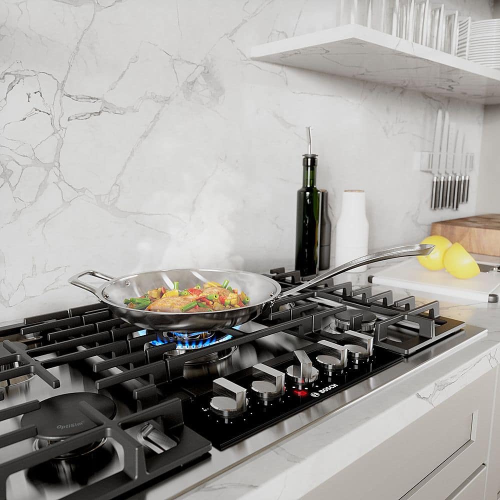 The image features a stove with a pan of food on it, and there are several spoons and knives nearby. The stove is part of a kitchen with a counter and a sink. The stove is made by Bosch, and it has a pan of food on it, indicating that someone is cooking. The presence of multiple spoons and knives suggests that the cook is well-equipped for preparing the meal.