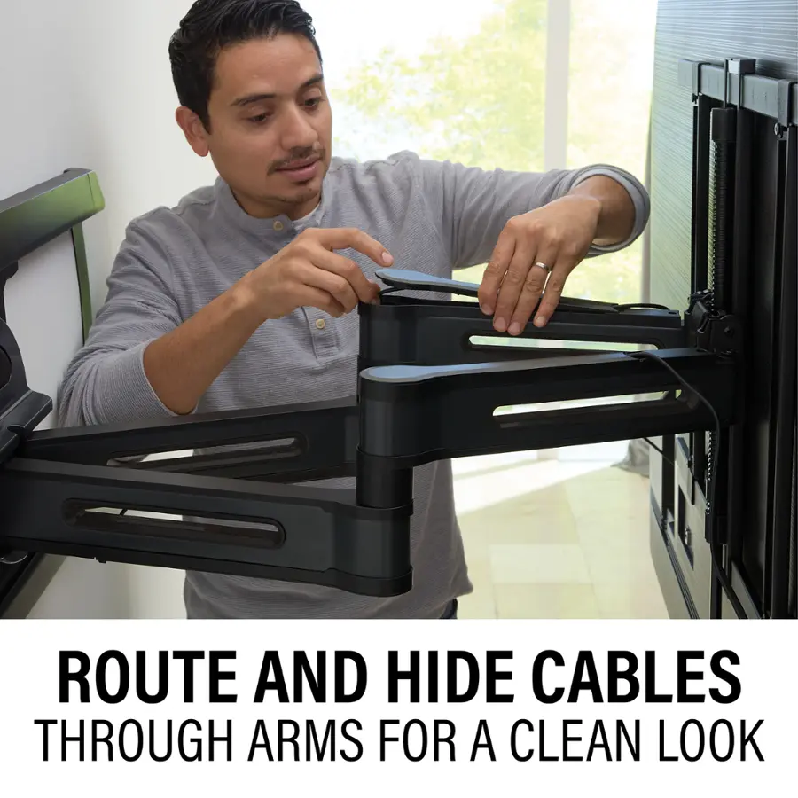 The image shows a man in a grey shirt attaching cables to a TV or a monitor. He is using a cable management system to route and hide the cables through the arms of the TV or monitor, creating a clean and organized look. This method of cable management is a practical and visually appealing way to keep cables organized and out of sight, ensuring a clutter-free and tidy appearance.